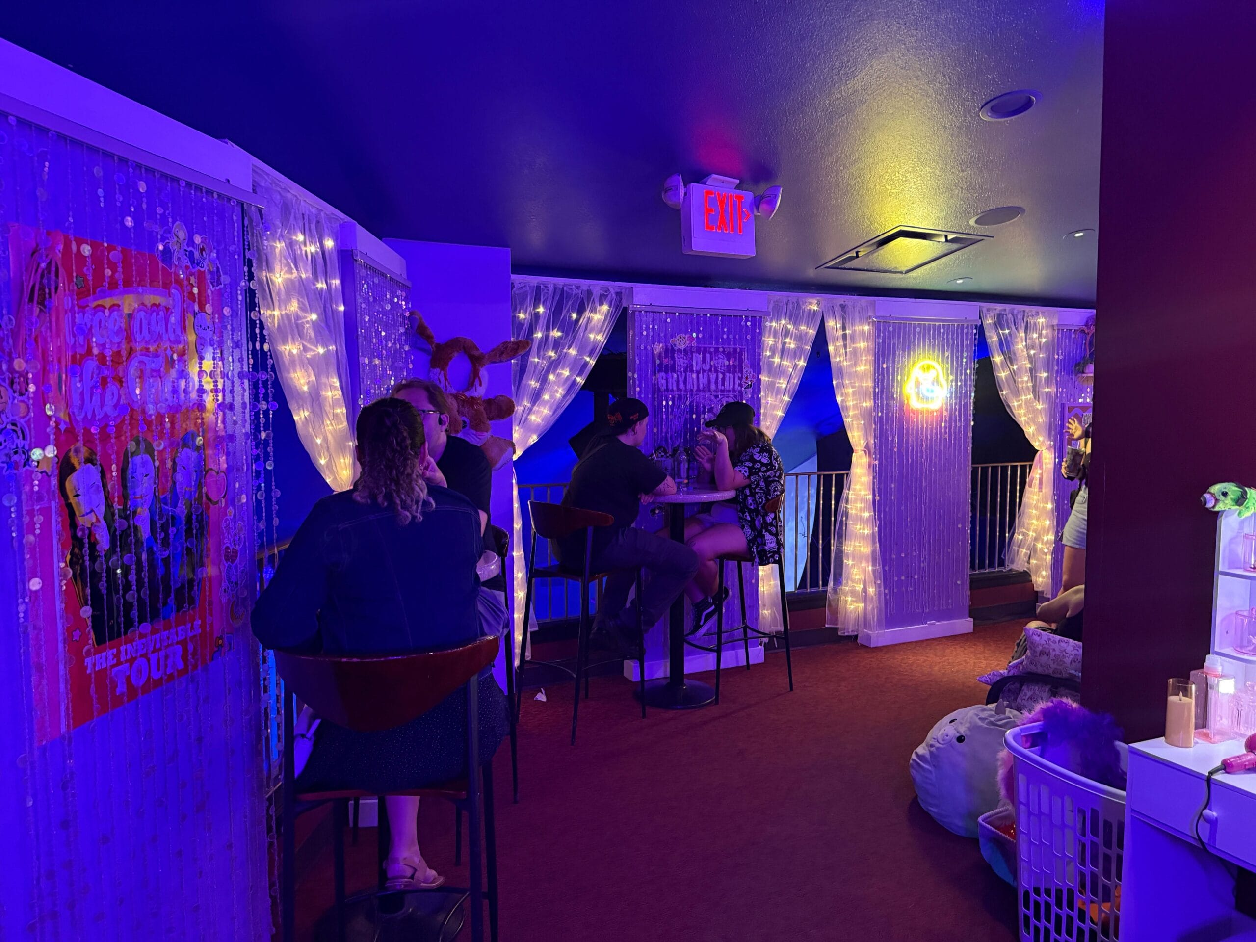 People relax at small tables under string lights at Dead Coconut Club, surrounded by purple lighting and themed posters.