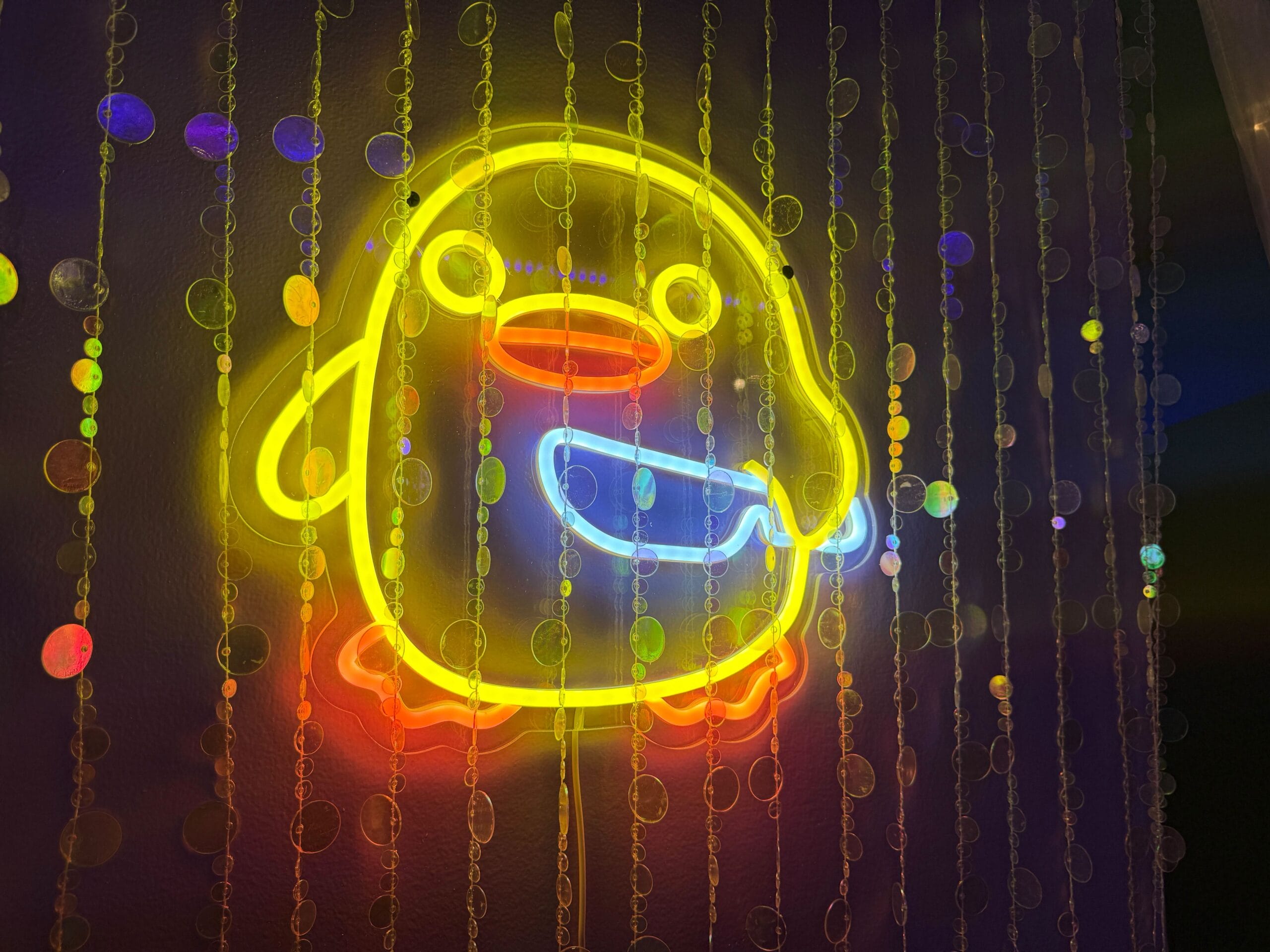 Neon yellow penguin sign glows at Dead Coconut Club behind clear beads on a dark purple wall.