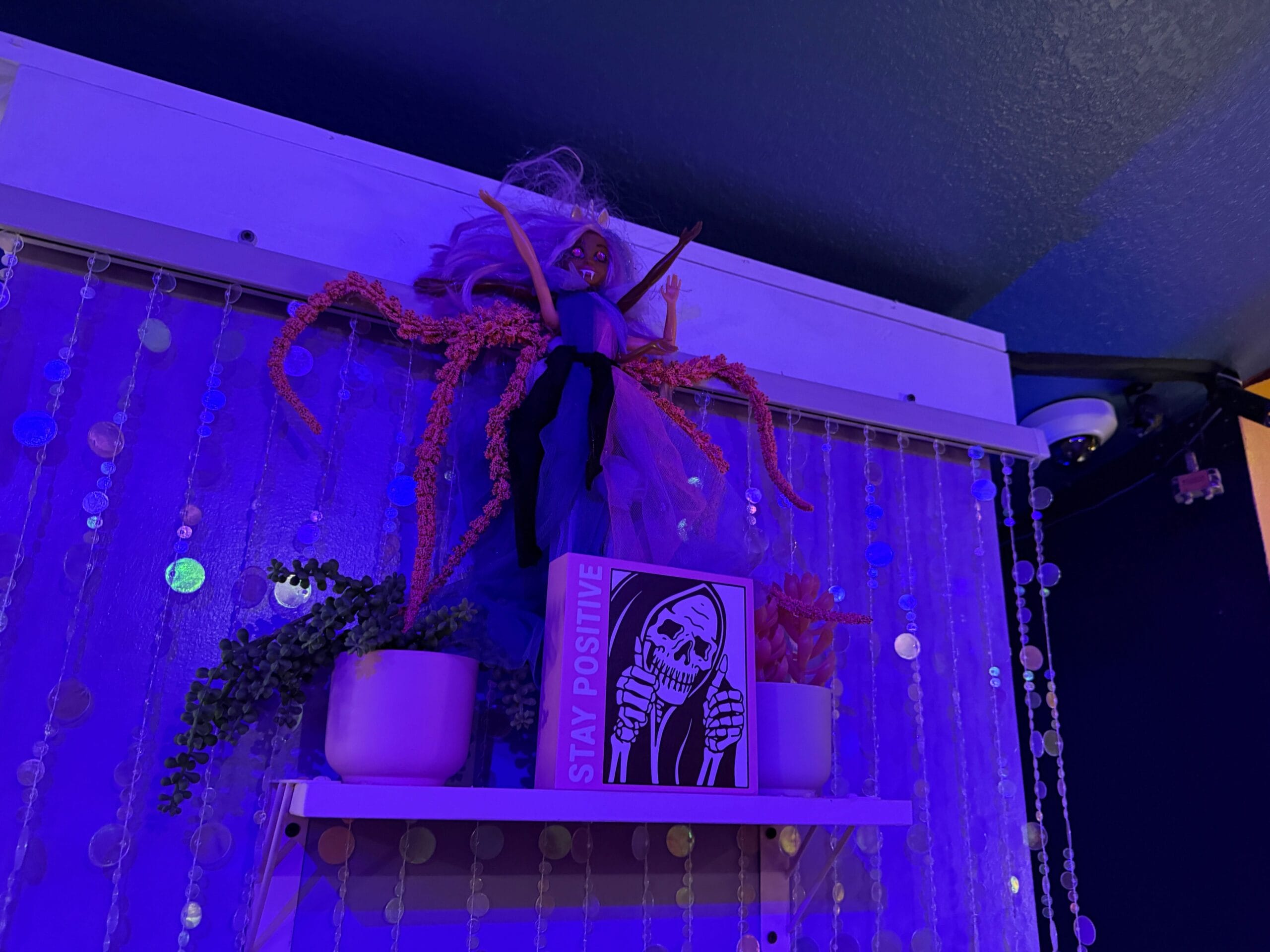 A Sorority Witches doll with outstretched arms stands near plants and a “Stay Positive” sign in purple lighting.