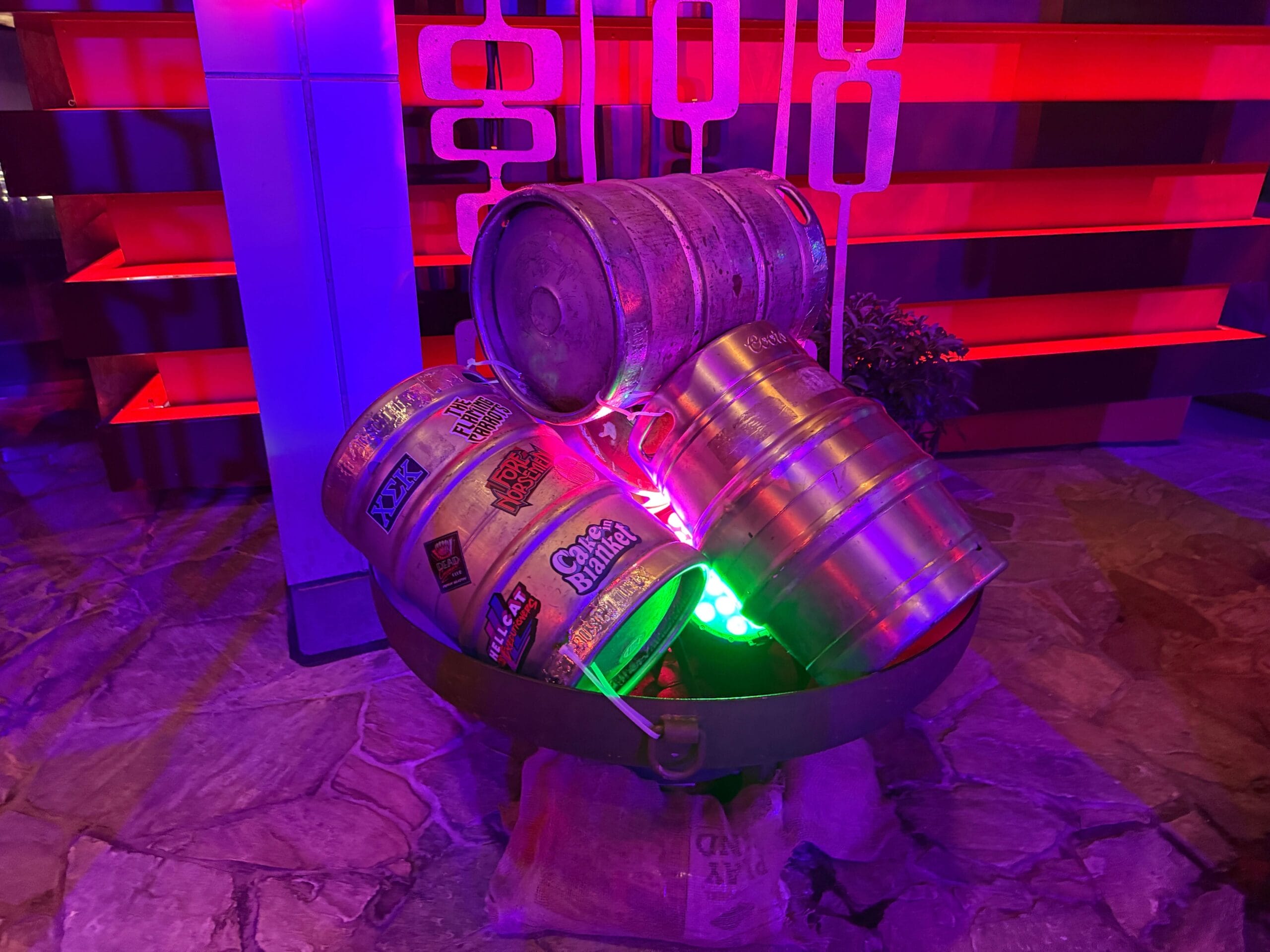 Three metal beer kegs with stickers sit stacked in a bowl, glowing in purple and green at Dead Coconut Club.