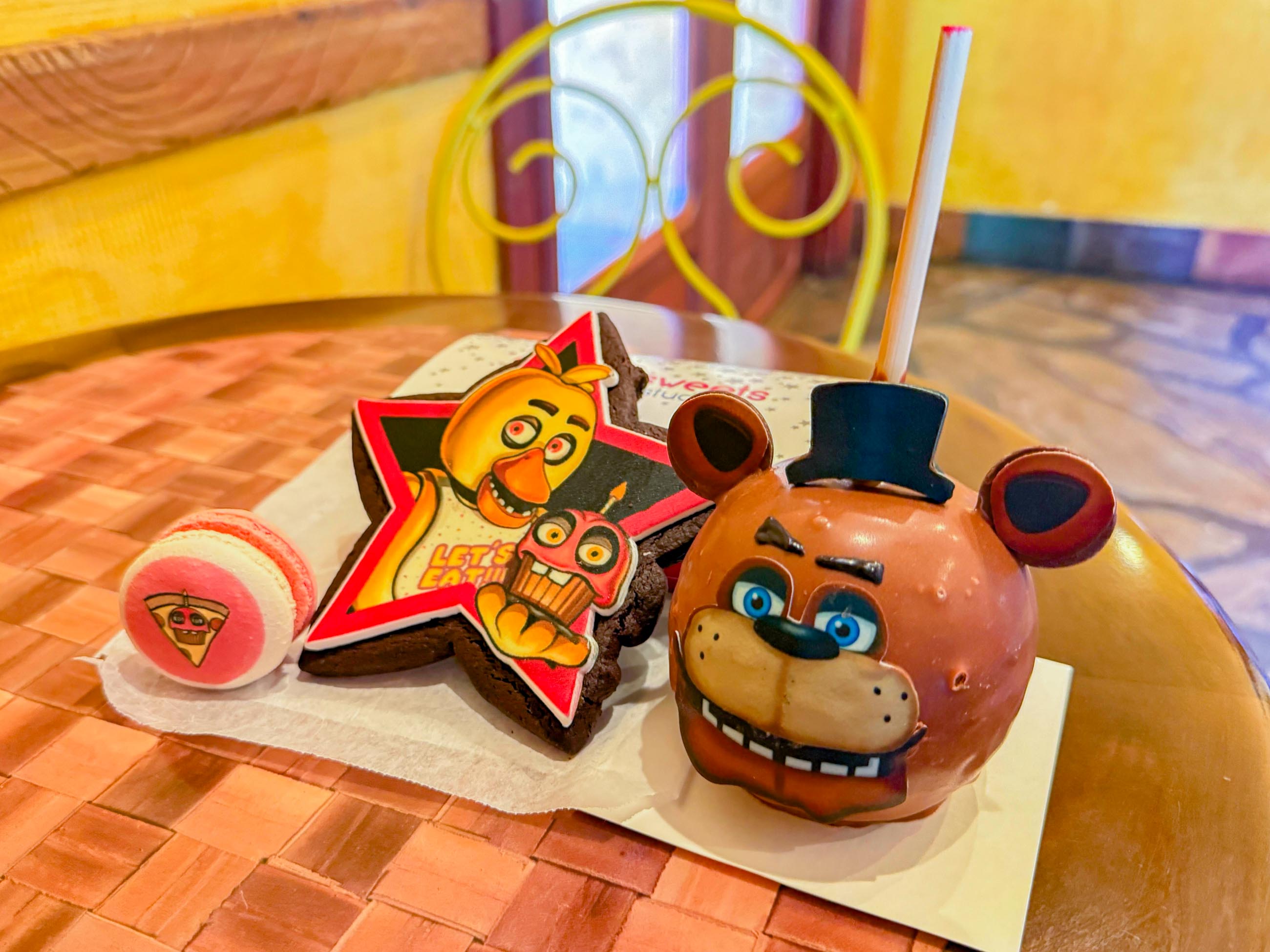 A macaron, star-shaped brownie with a cartoon bird, and Five Nights at Freddy's bear-shaped chocolate apple are on a woven table.