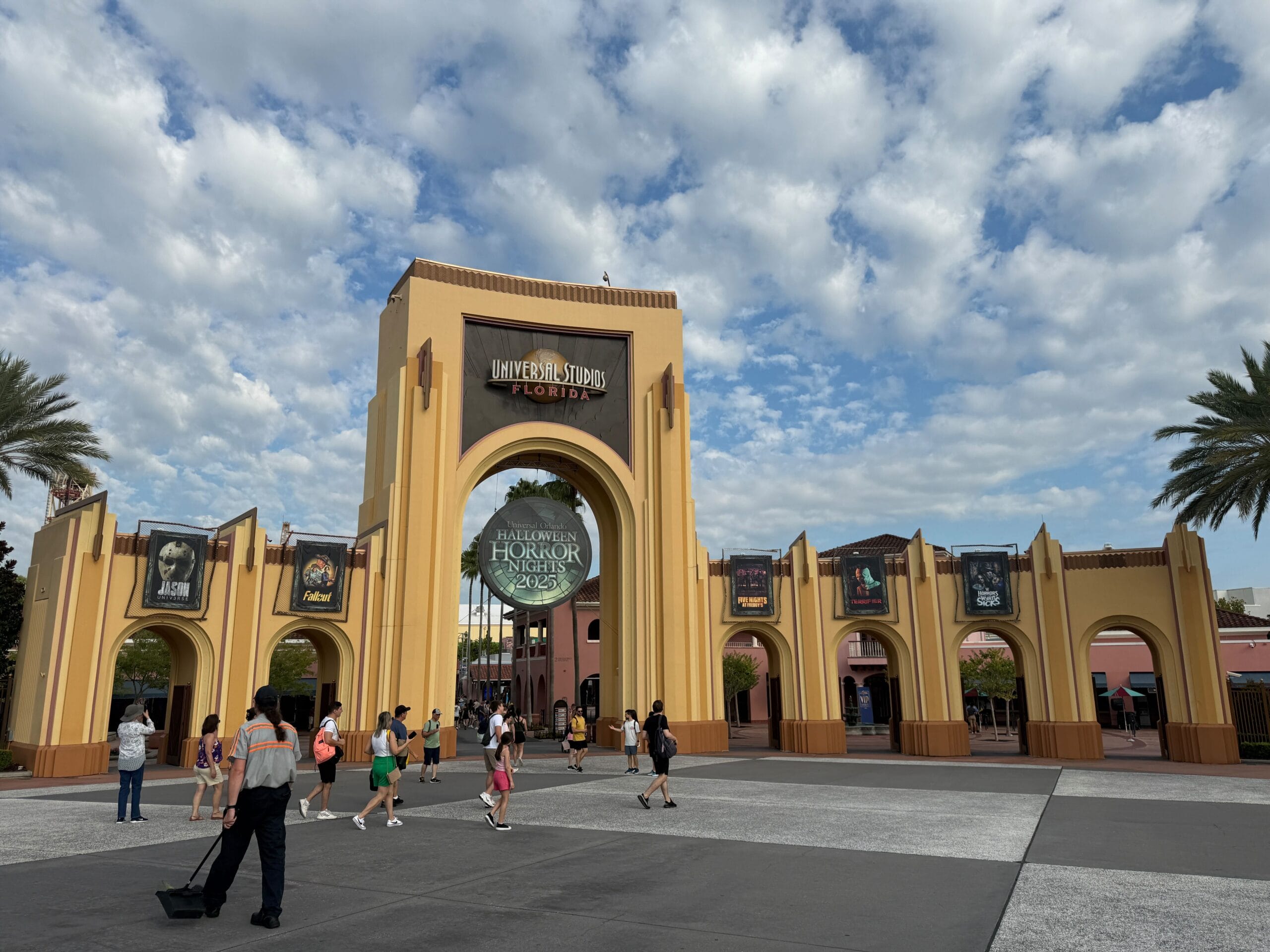 Entrance to Universal Studios Florida with Halloween Horror Nights banners and people walking under a partly cloudy sky.
