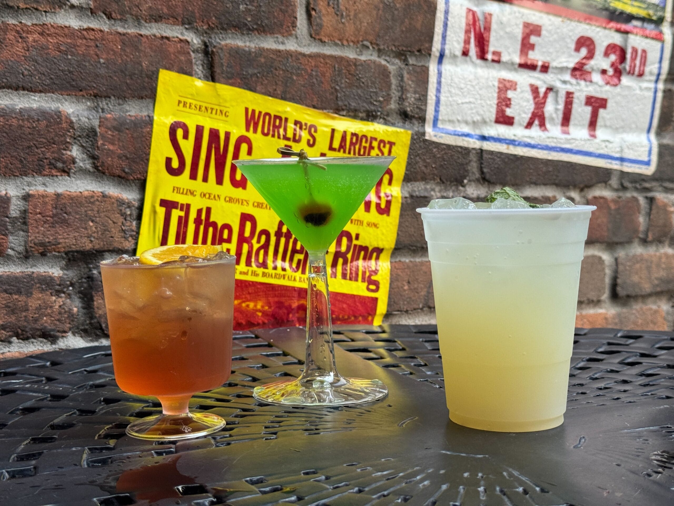 Three colorful cocktails on a black outdoor table at Black Cat Lounge, in front of a brick wall with vintage signs.