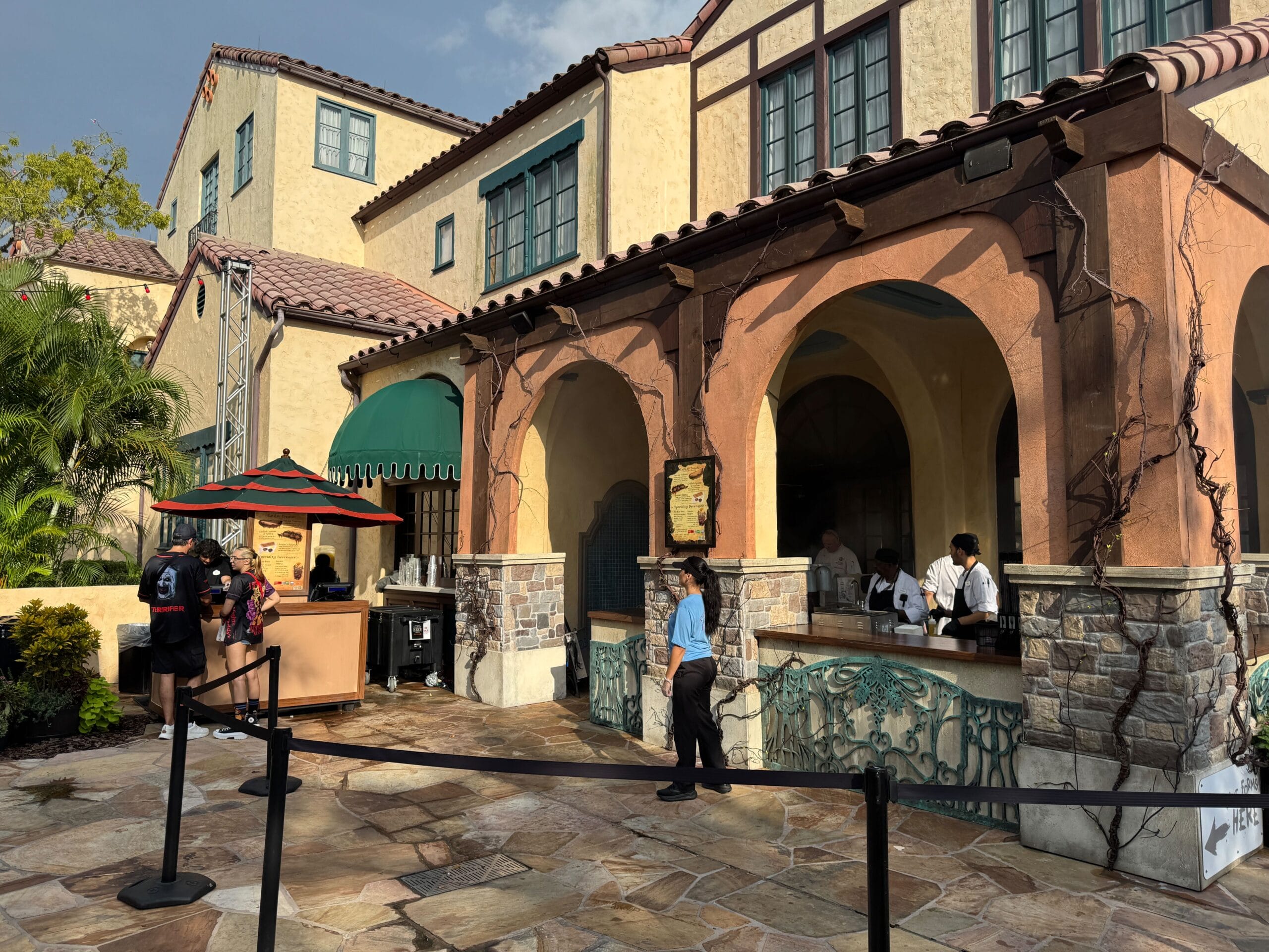 People order food at the Eats & Treats outdoor counter of a Mediterranean-style restaurant with stone and stucco walls.