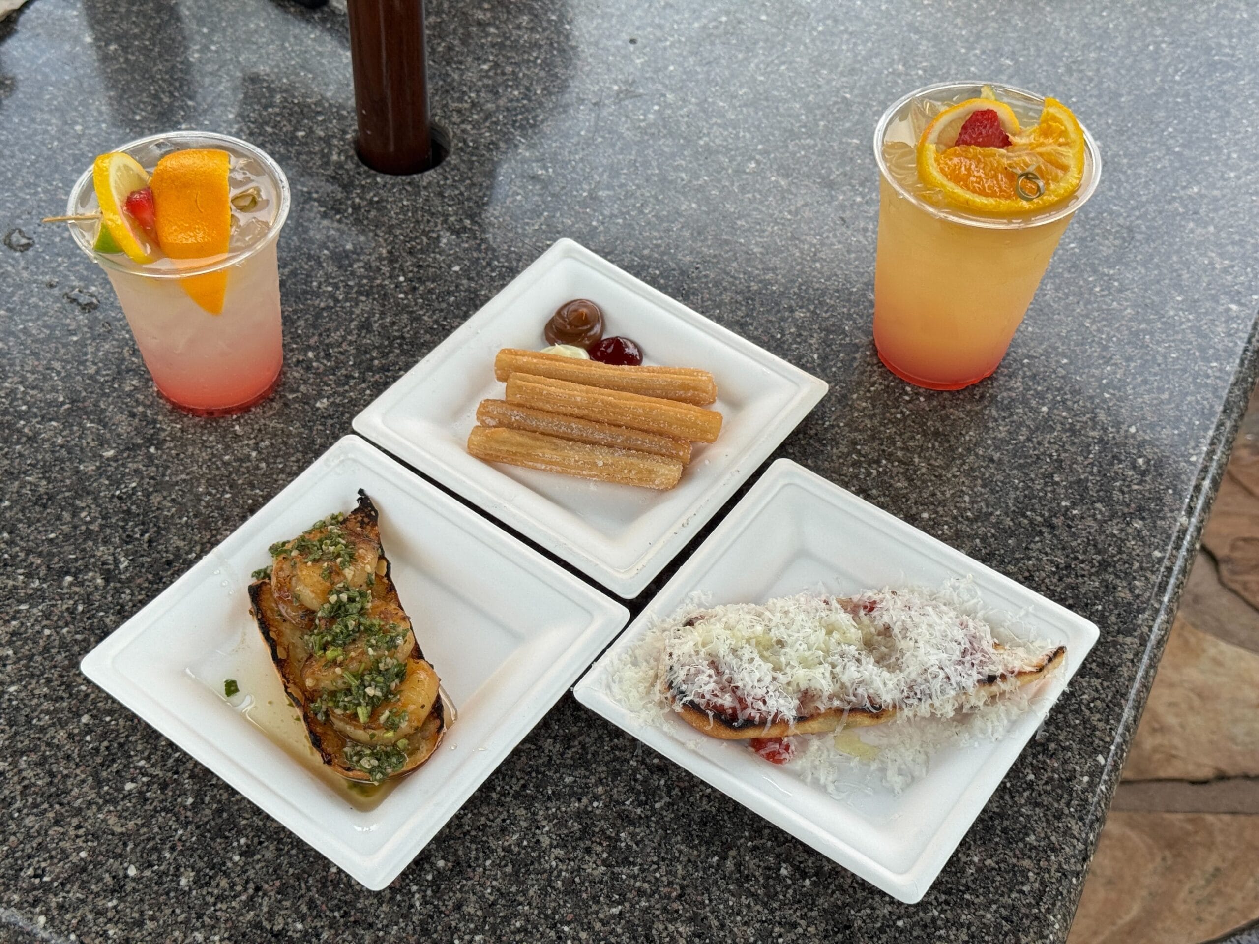 Two fruity drinks and three plates of Eats & Treats at El Artista on a stone table: churros, cheesy toast, roasted eggplant.