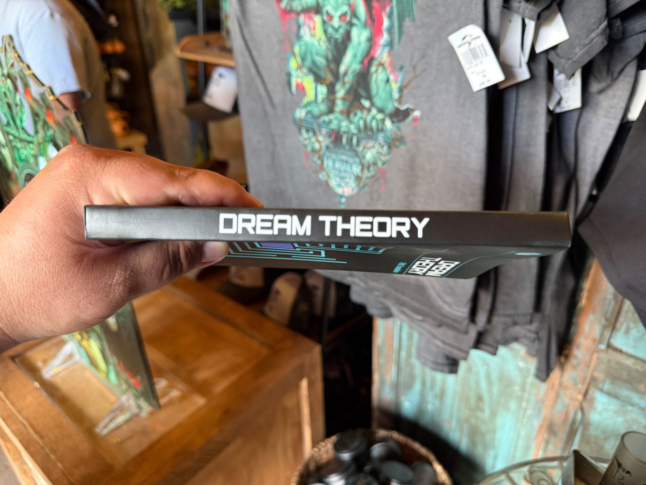 A hand holds a "DREAM THEORY" book with Halloween Horror Nights 34 T-shirts displayed in the background.