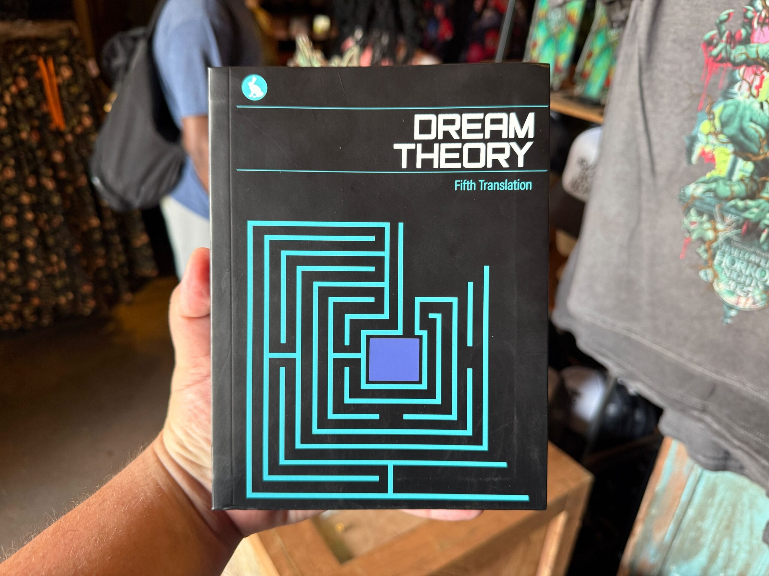 A hand holds a book titled "Dream Theory: Fifth Translation" beside a Firefly Fun House Sign with a maze design.