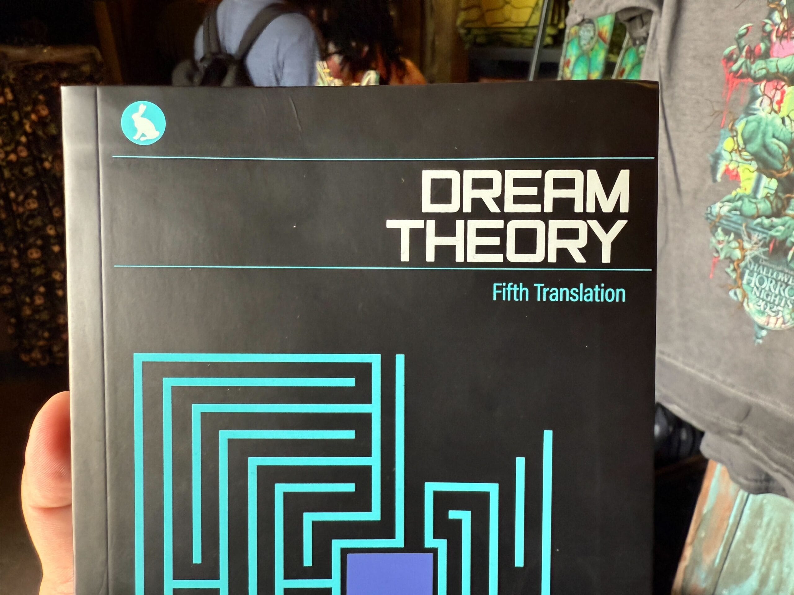 A hand holds a book titled "DREAM THEORY Fifth Translation" with a geometric blue pattern, spotted at Halloween Horror Nights 34.