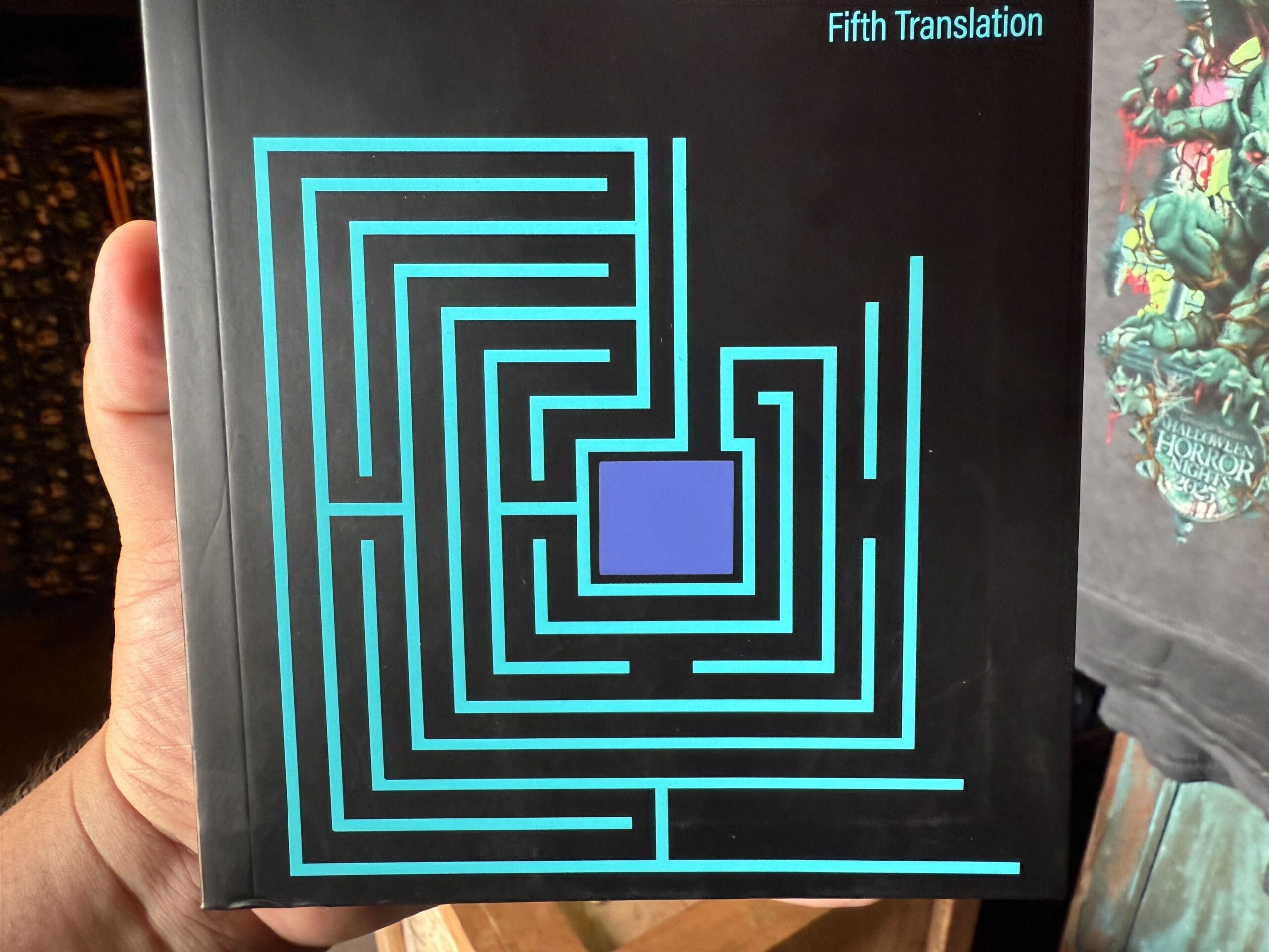 A hand holds a "Fifth Translation" book with a maze cover, beside a Clown Cafe Halloween Horror Nights 34 display.
