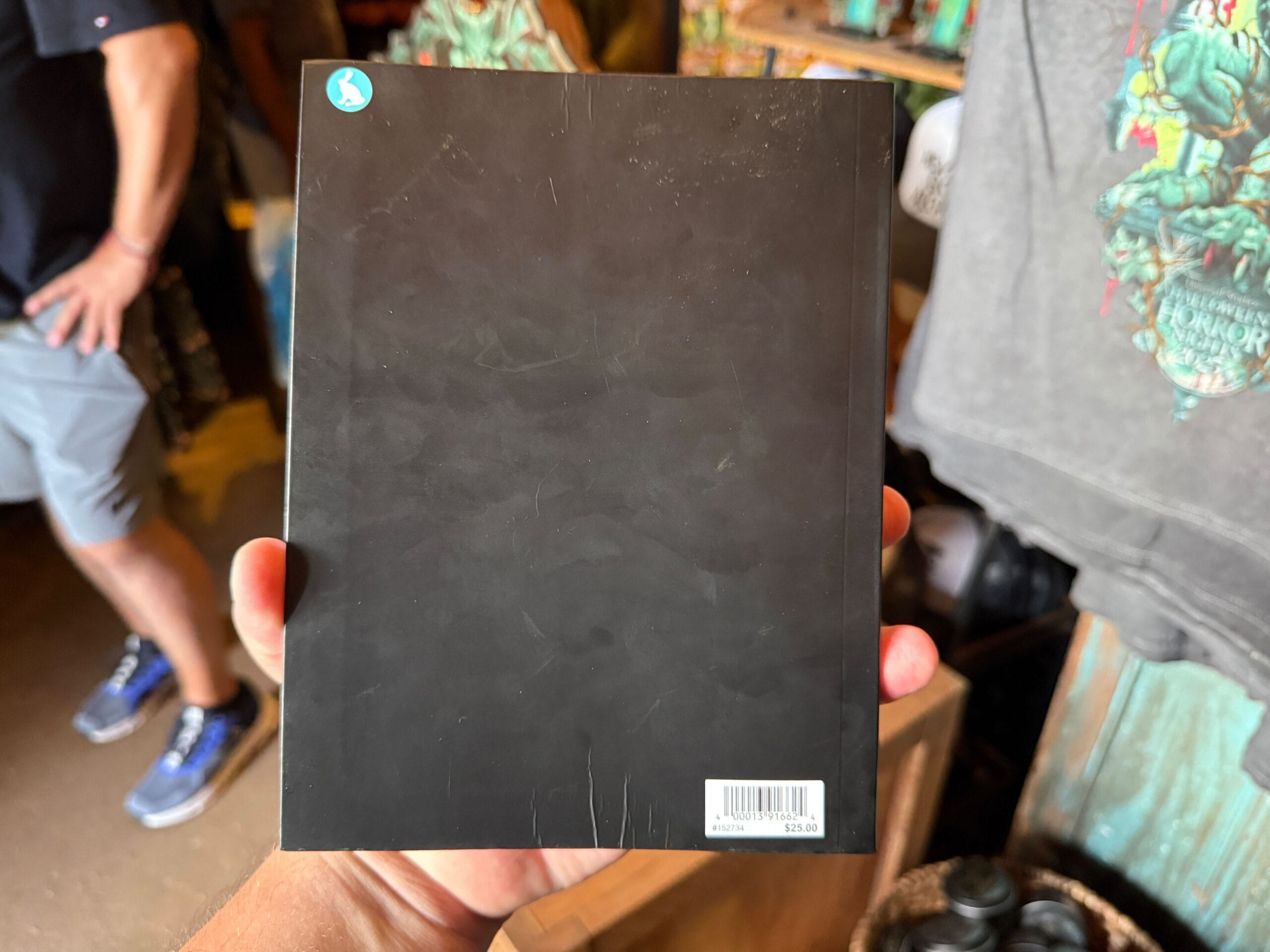 A person holds a plain black book with a barcode; Halloween Horror Nights 34 merchandise is visible in the background.