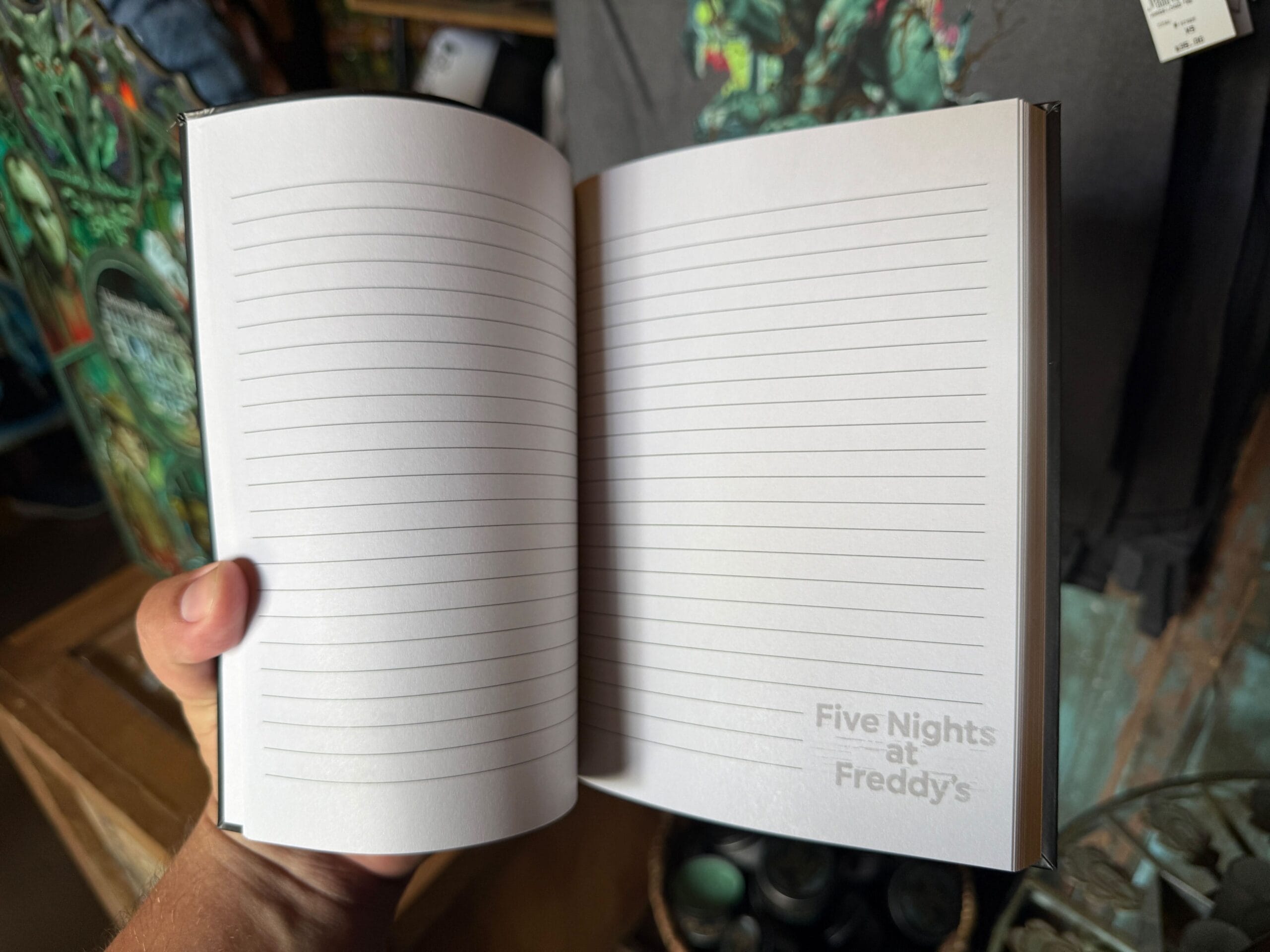 A hand holds open a lined notebook featuring the "Five Nights at Freddy's" logo and a Clown Cafe sketch in the corner.