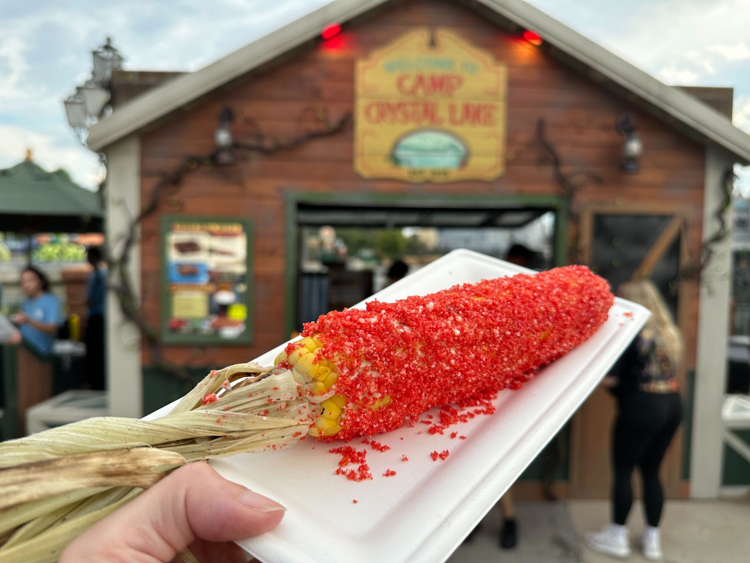 A hand holds a corn on the cob coated in red seasoning at the Camp Crystal Lake food stand—perfect for Halloween Horror Nights.