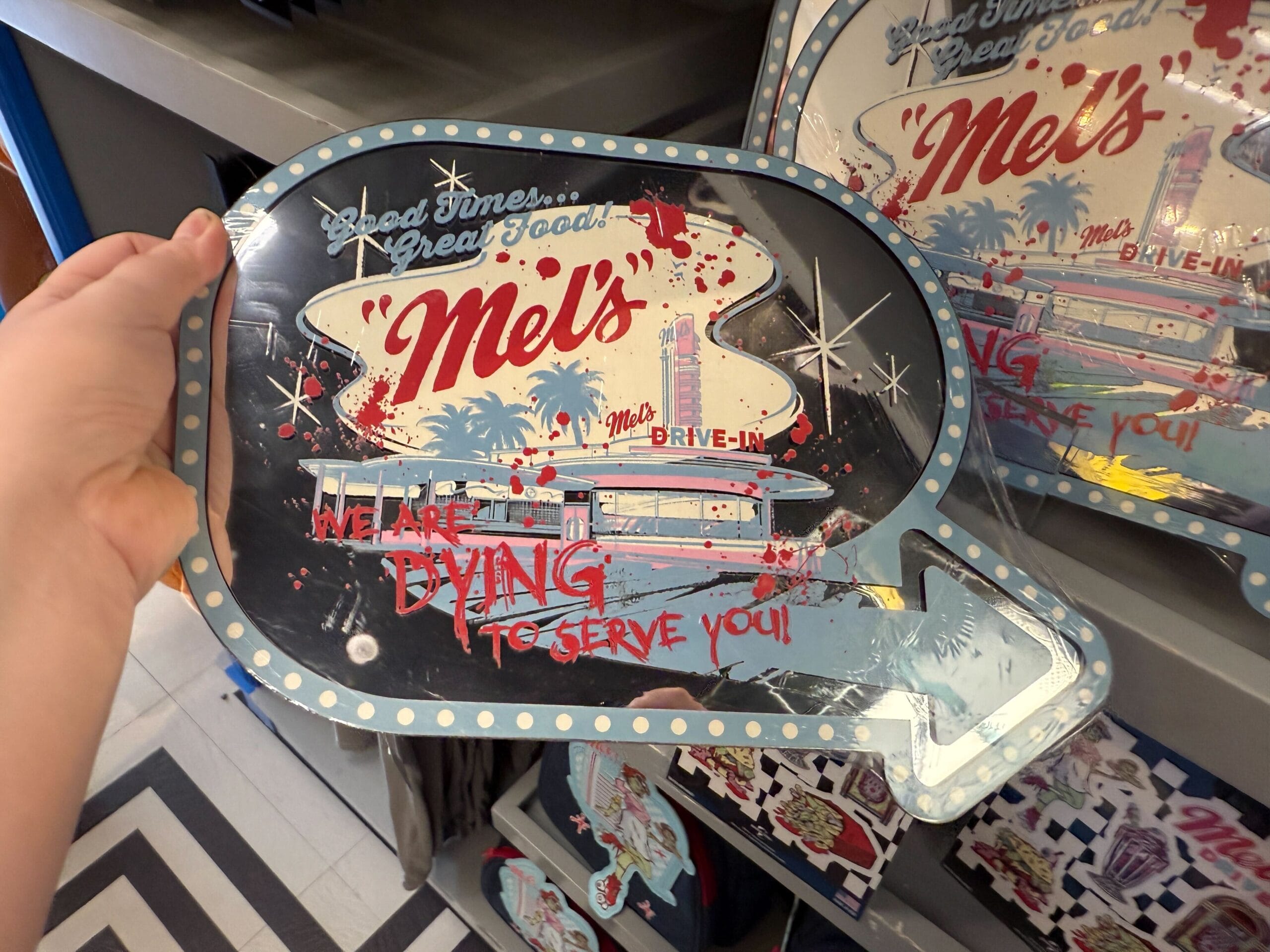 A hand holds a retro-style Mel's Die-In sign that says, "We are dying to serve you! Zombies merch, anyone?