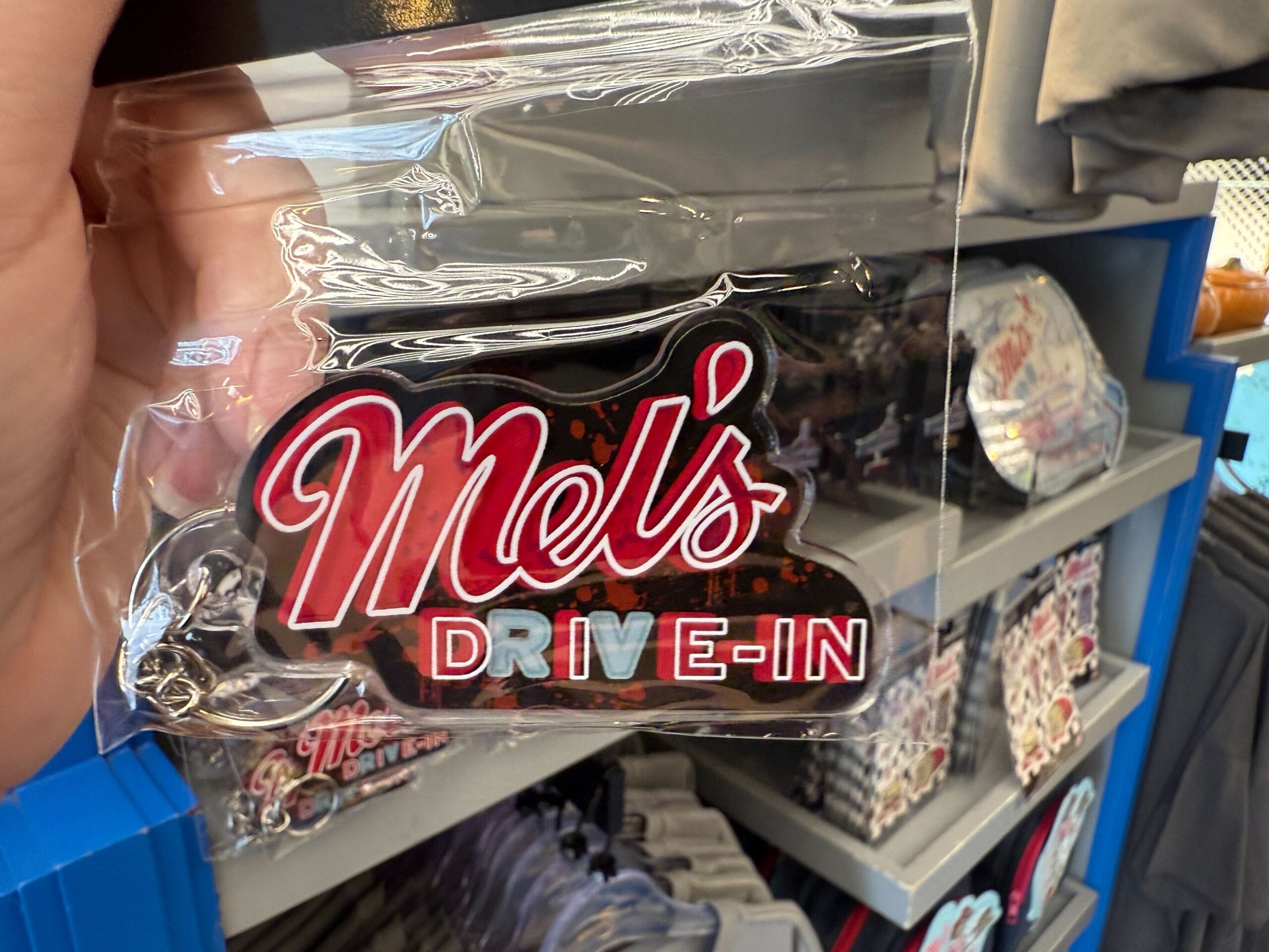 A hand holds a “Mel’s Drive-In” keychain in front of Mel's Merch display with more keychains and shirts.