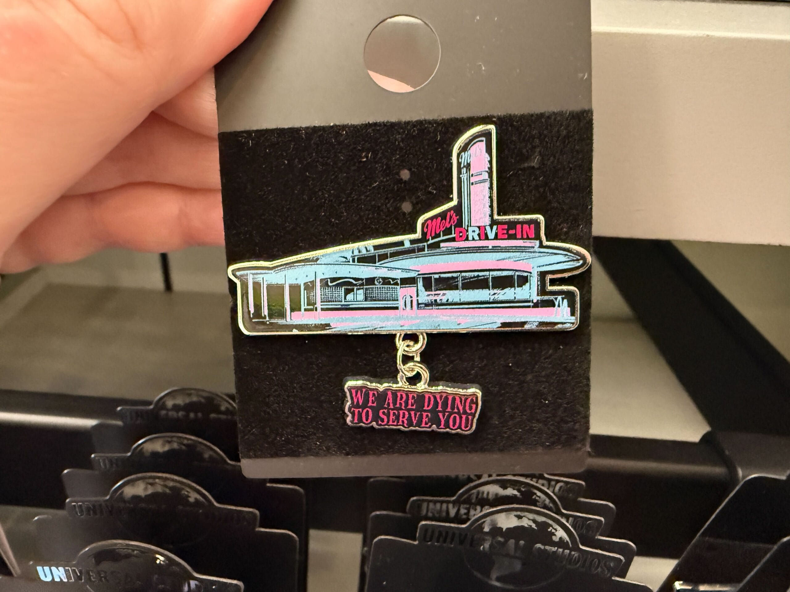 A hand holds a Die-In Zombies Merch pin of a vintage diner labeled "Mel's Drive-In" with "WE ARE DYING TO SERVE YOU.