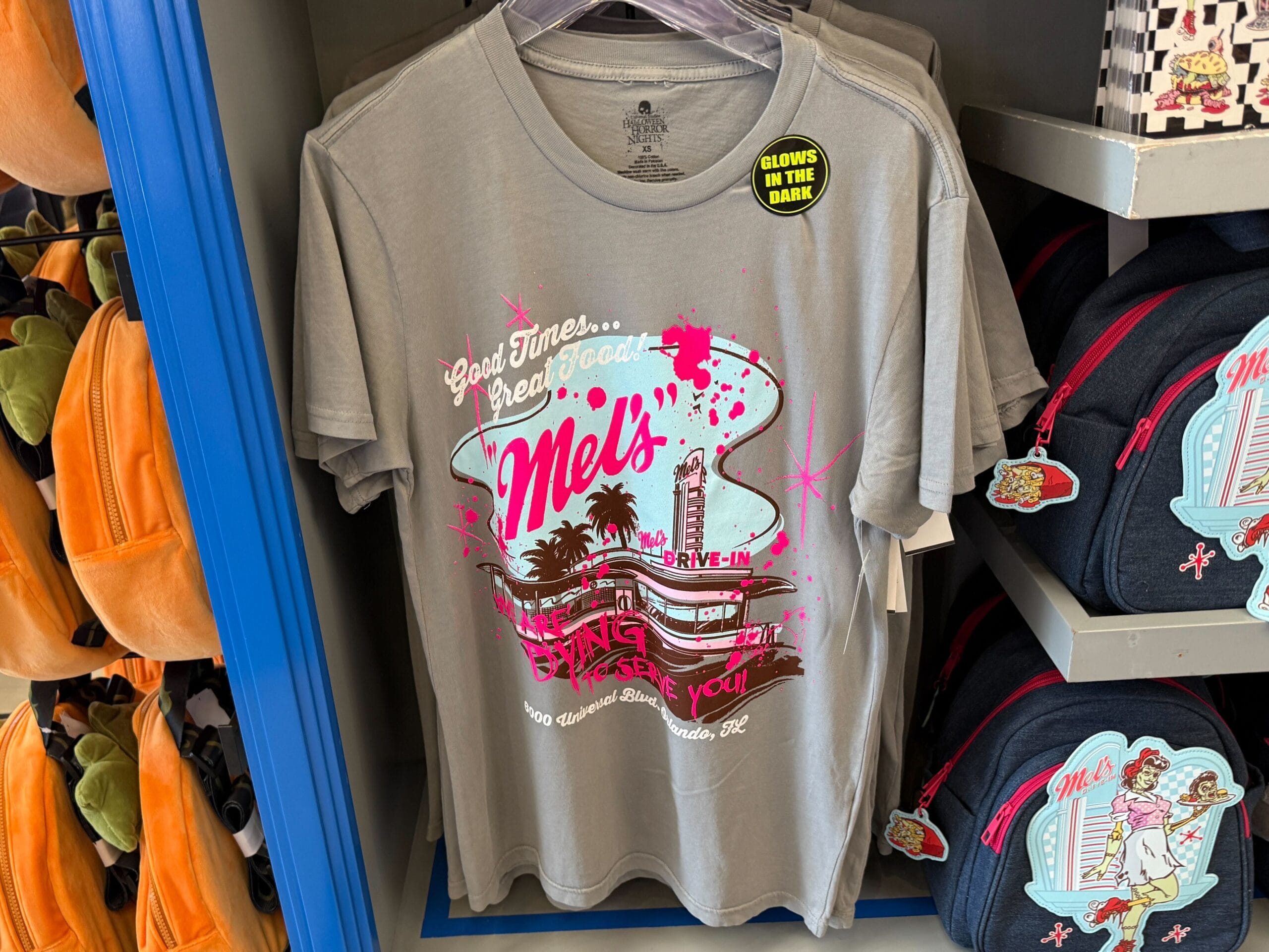 Gray t-shirt with a retro "Mel's Die-In" graphic, neon lights, palm trees, and a "Glows in the Dark" sticker—zombie merchandise.