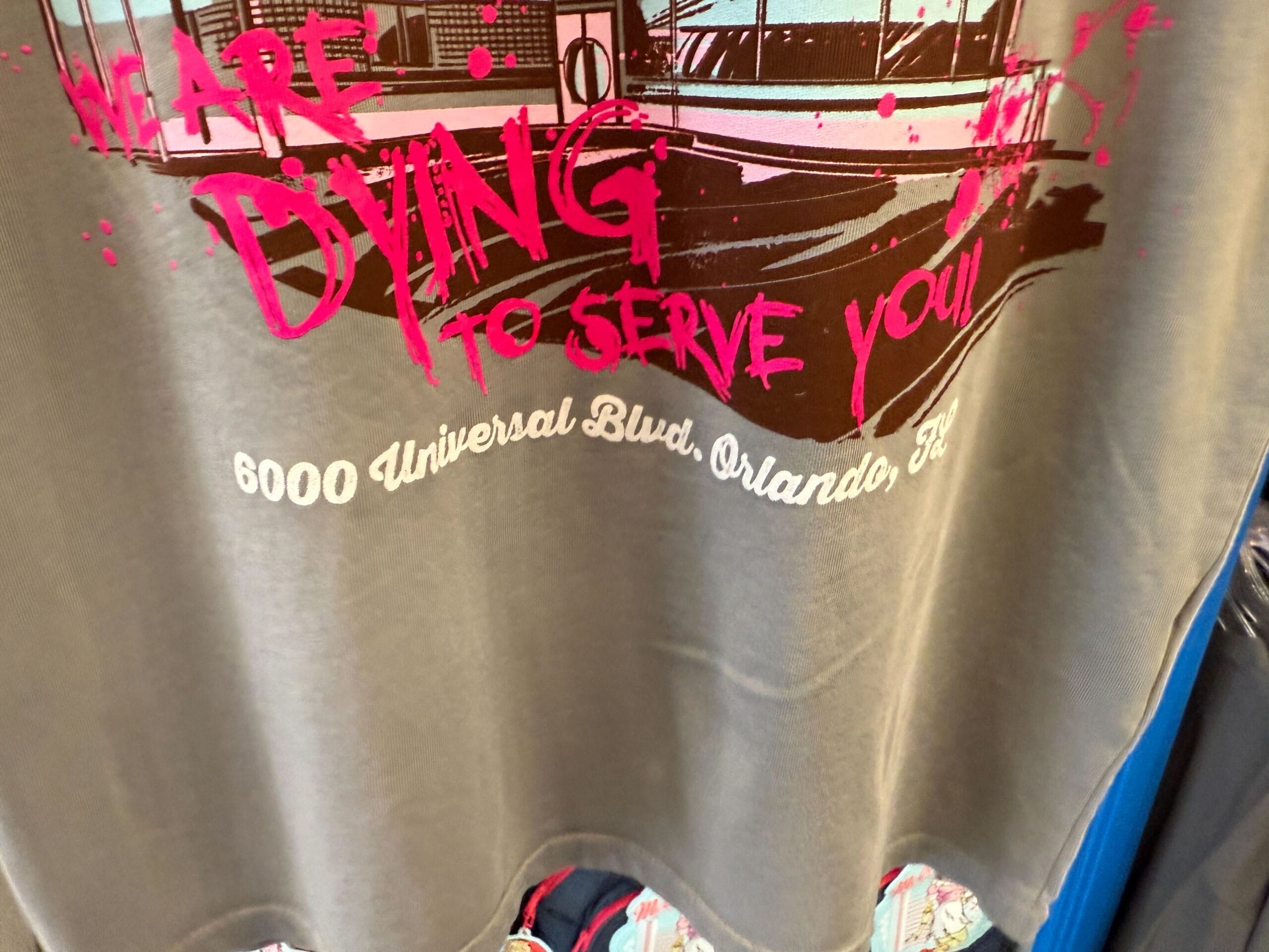 Gray shirt featuring "WE ARE DYING TO SERVE YOU!" Die-In Merch, 6000 Universal Blvd, Orlando, FL in pink and white text.