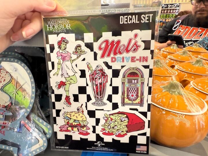 Hand holds a "Mel's Die-In" Halloween Horror Nights decal set with Die-In Zombies diner graphics.