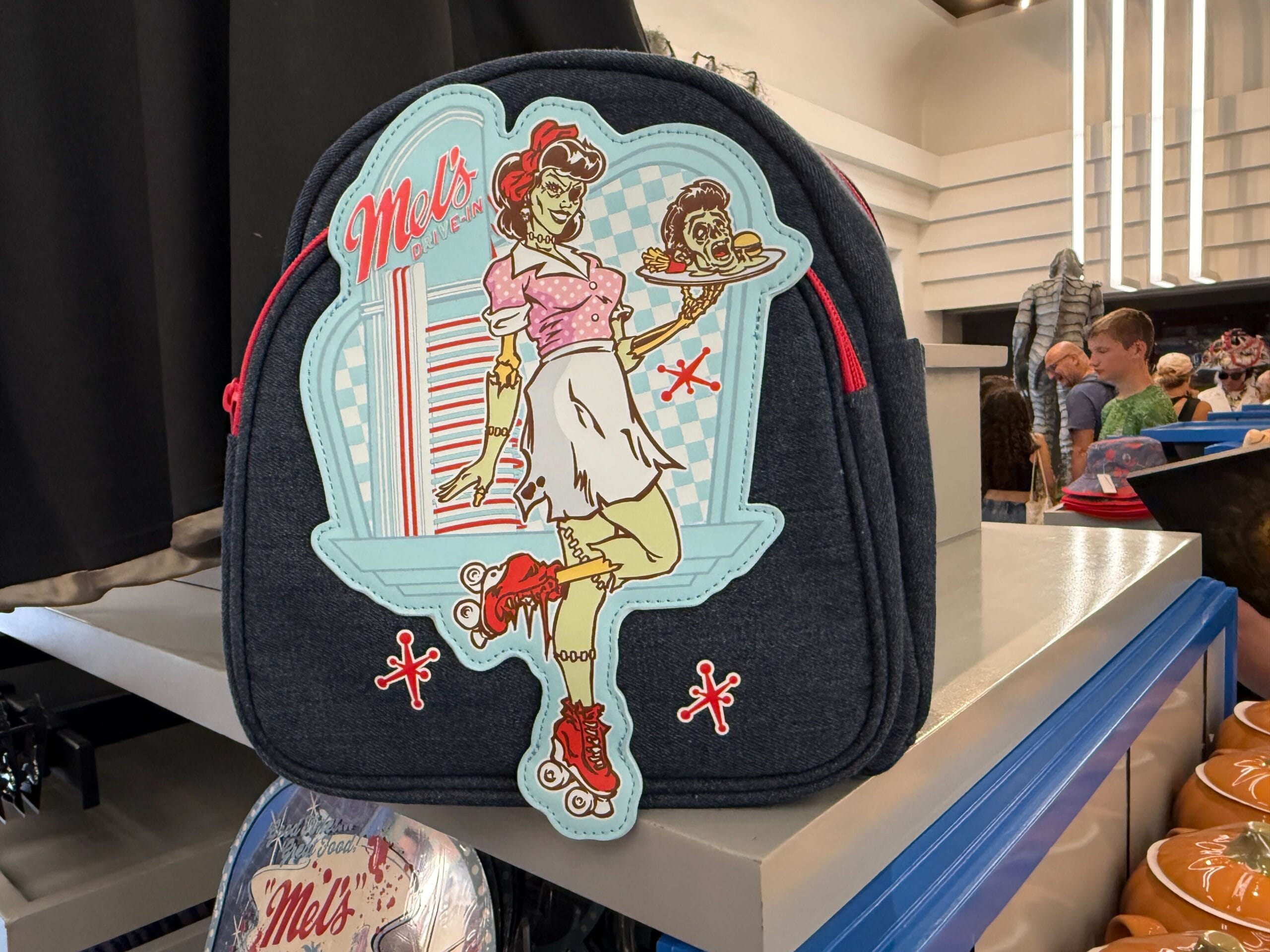 A denim backpack with a retro diner waitress illustration, labeled "Mel's," joins the latest Mel's Merch on the store shelf.
