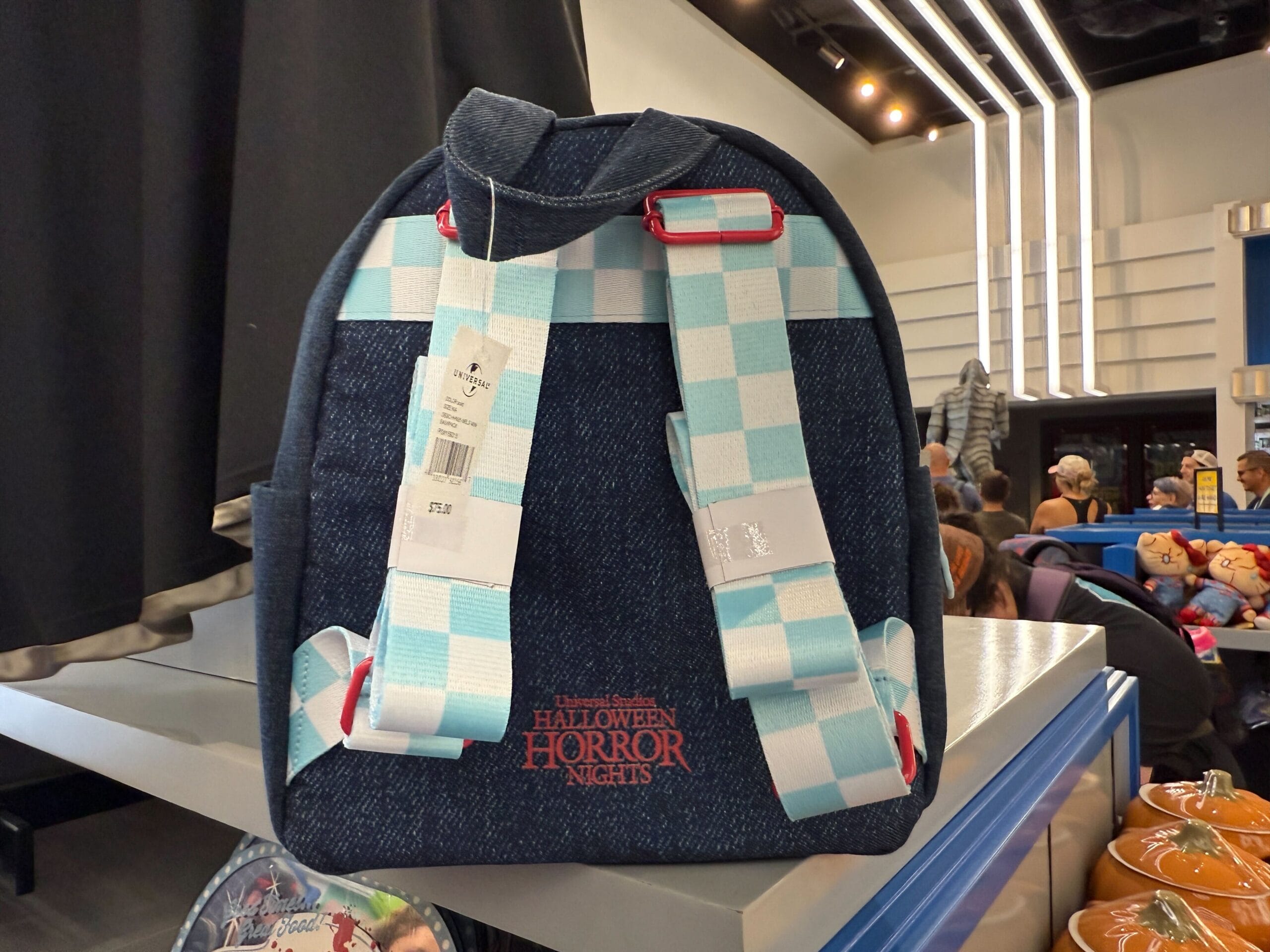 A blue backpack with white checkered straps, "Halloween Horror Nights" logo, and exclusive Zombies merch displayed in a store.