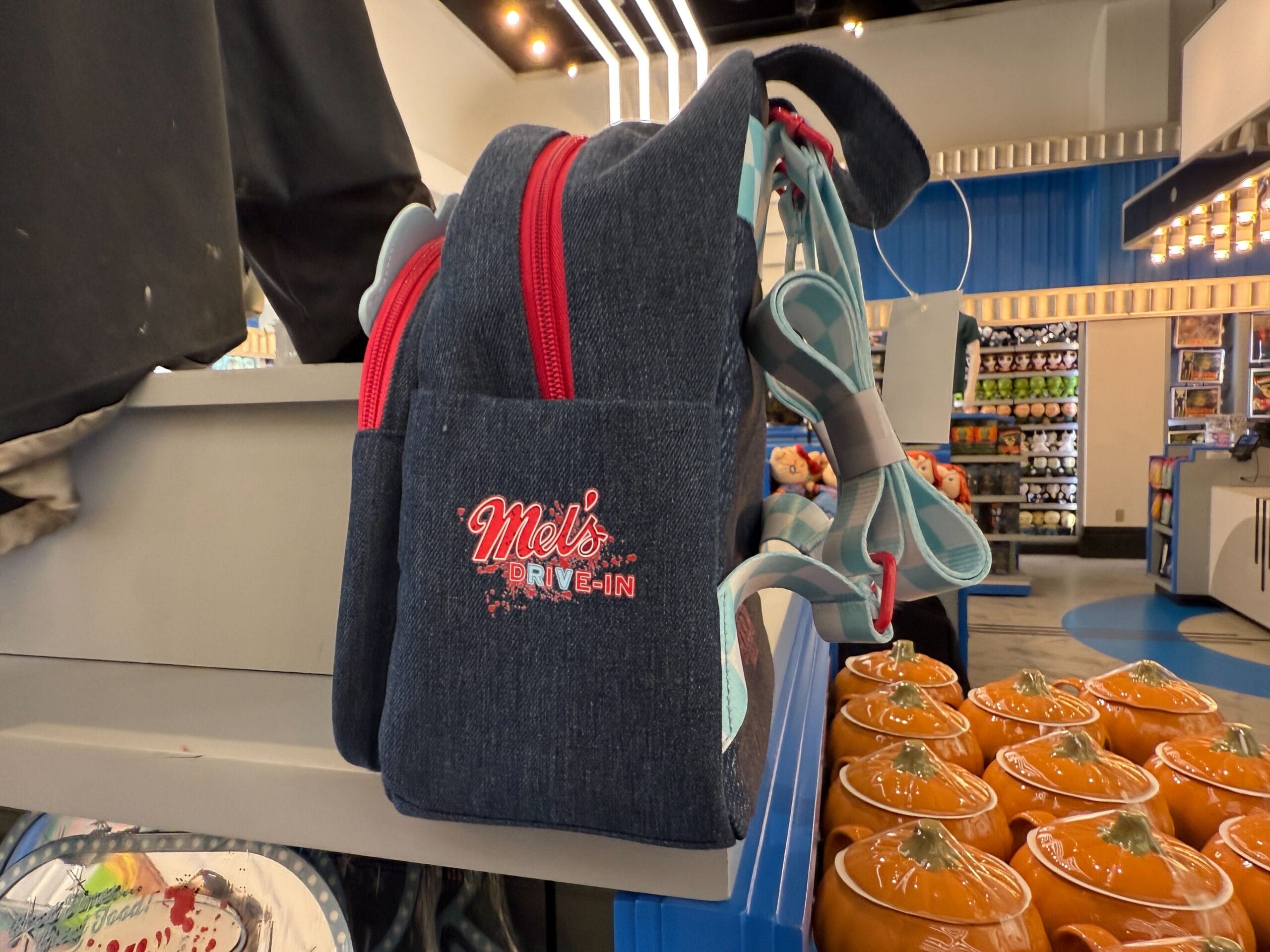 A blue denim Merch backpack with "Mel's Drive-In" logo sits on a shelf near pumpkin-shaped containers.