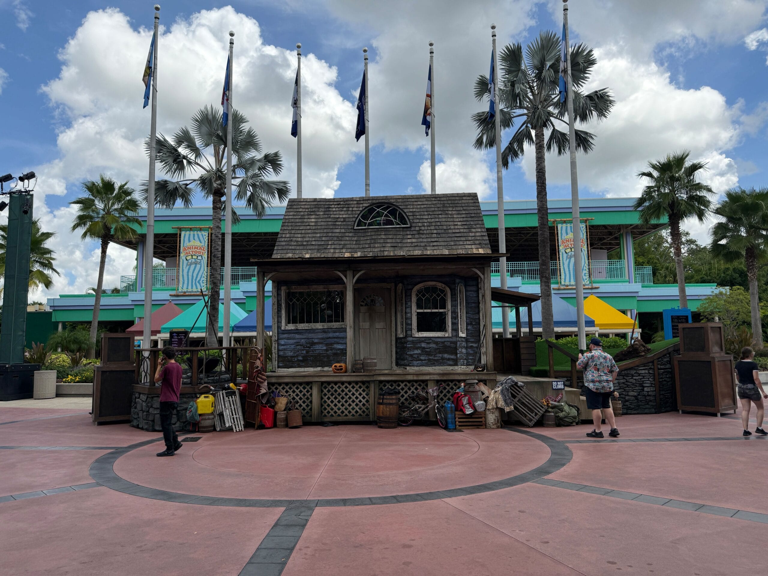 A small rustic wooden house on stage with a Firefly Fun House Sign, palm trees, and people walking nearby.