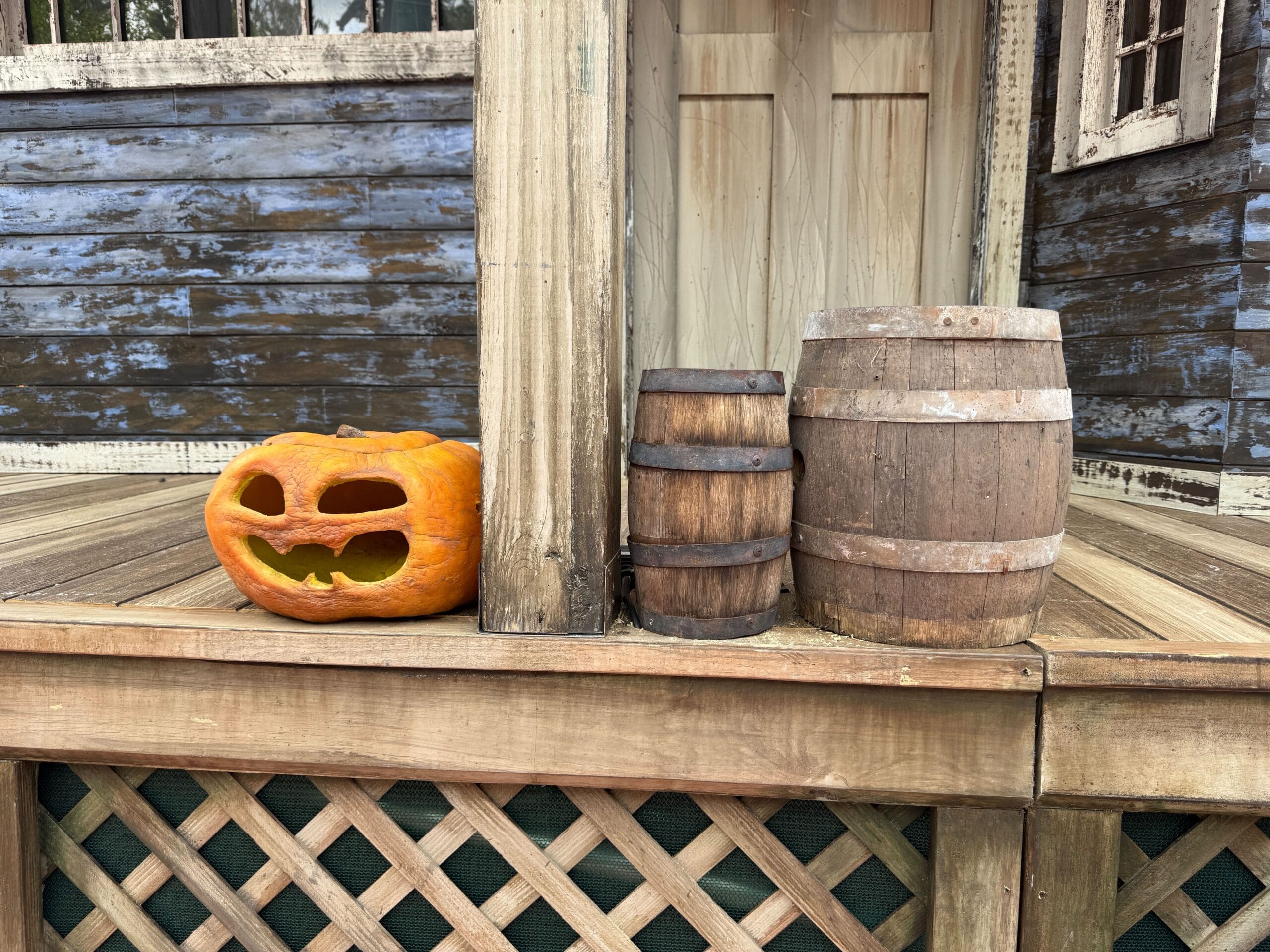 Carved pumpkin and two wooden barrels on a porch, evoking Clown Cafe vibes with a weathered blue house in the background.
