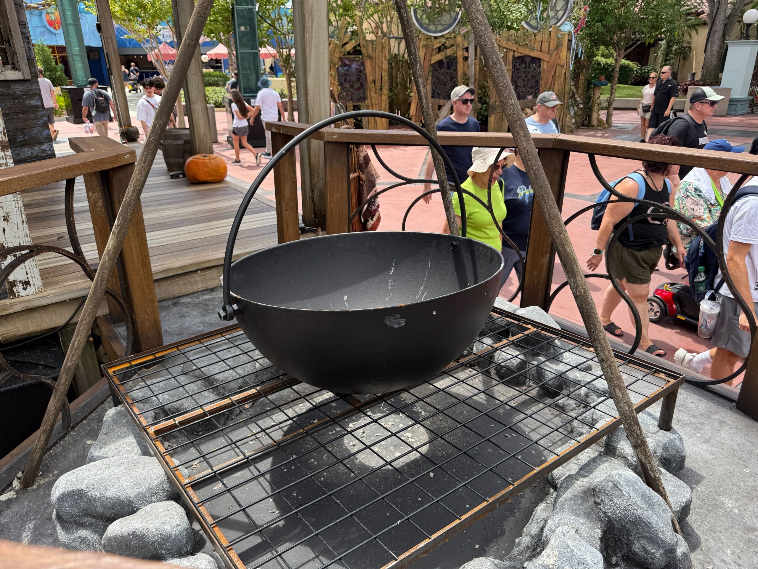 A large black cauldron sits on a metal grate at Halloween Horror Nights 34, with people walking in the background.