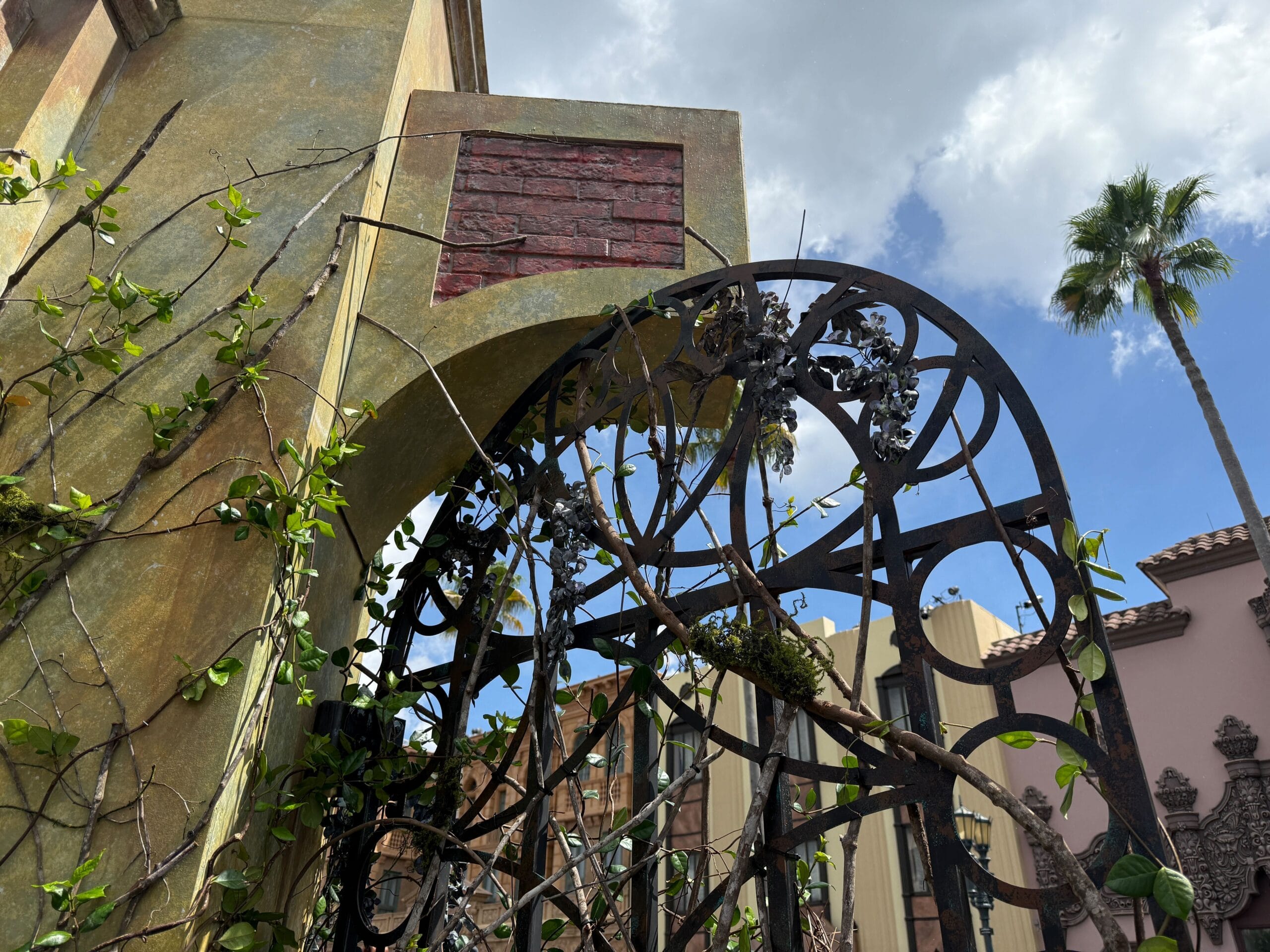 Wrought iron gate with vines under an archway, blue sky, clouds, and a Clown Cafe sign in the background.