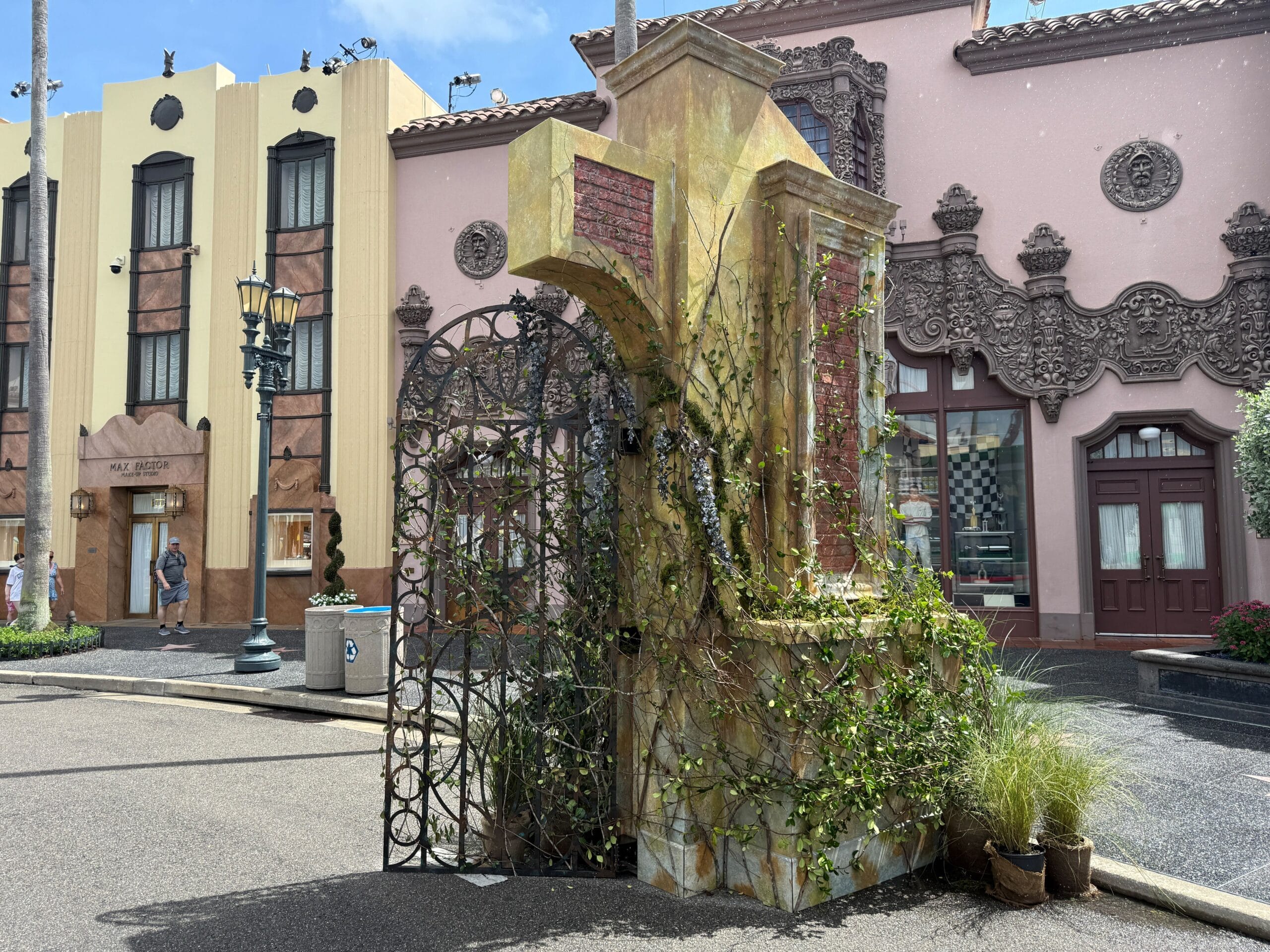 A large weathered cross with vines stands near ornate buildings by the Clown Cafe on a sunny street.