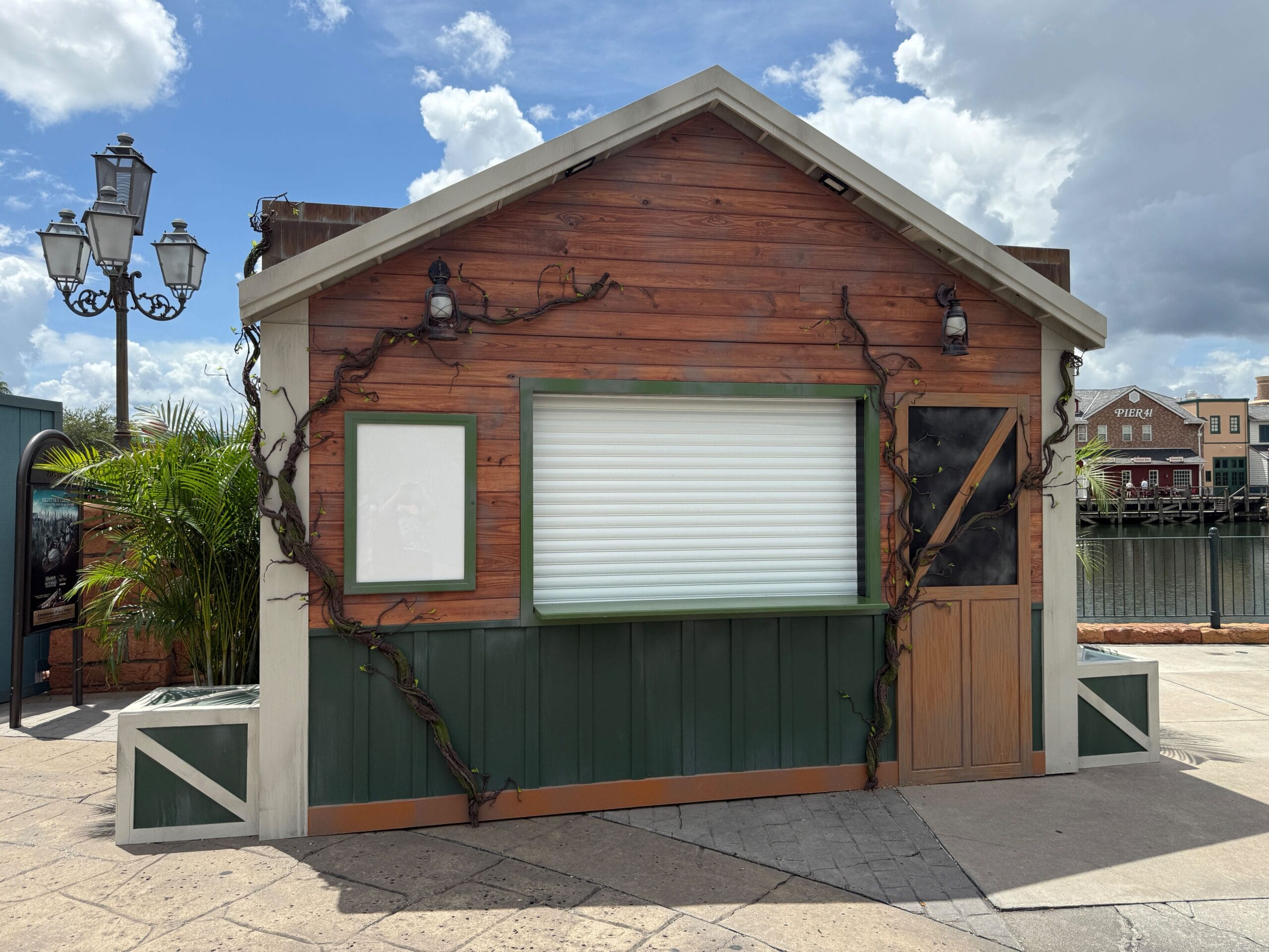 A small wooden kiosk with a closed window, vine decor, and a Firefly Fun House Sign under a partly cloudy sky.