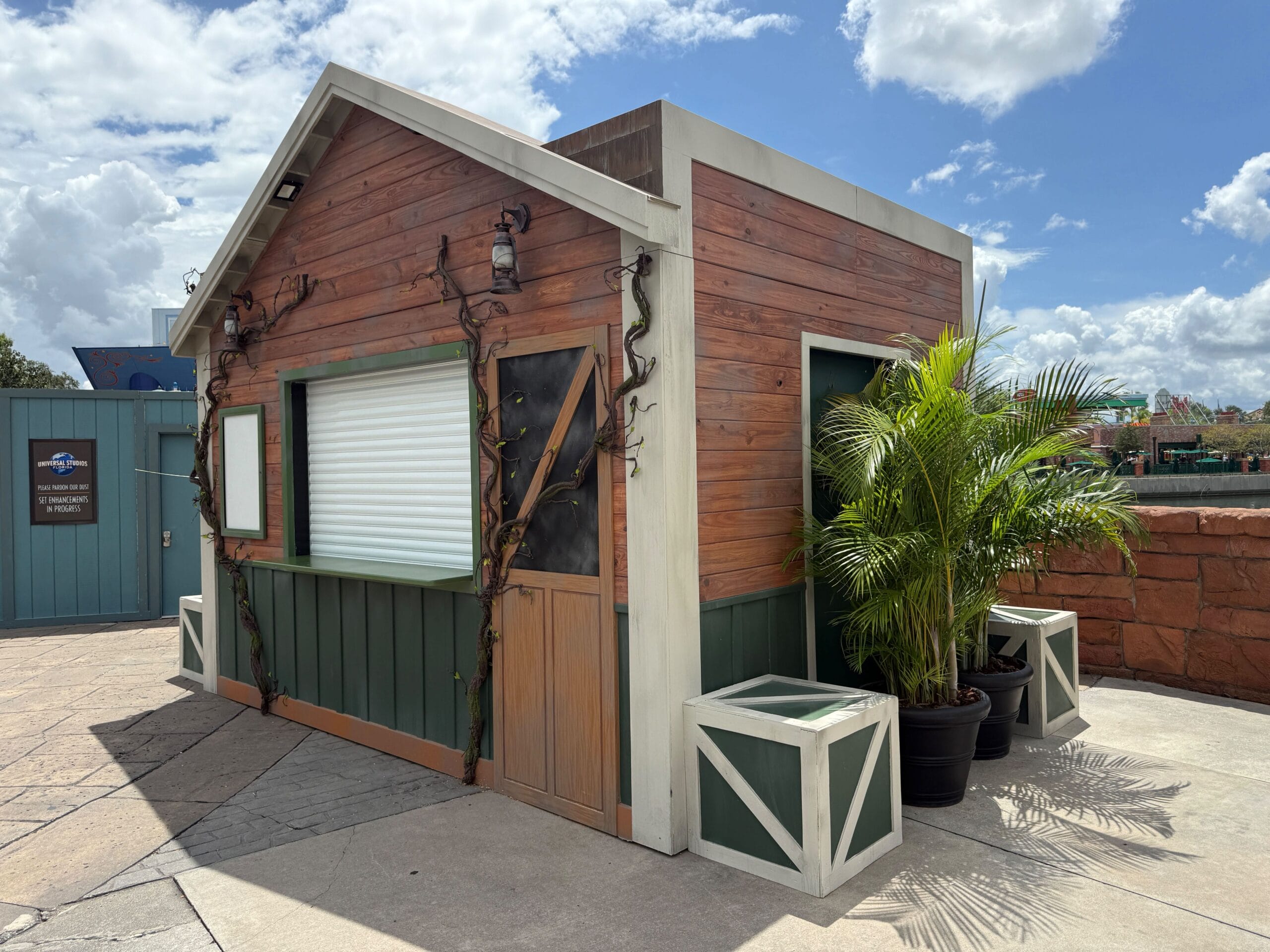 Small wooden kiosk with potted plants, closed shutters, and a Firefly Fun House Sign under a cloudy sky.