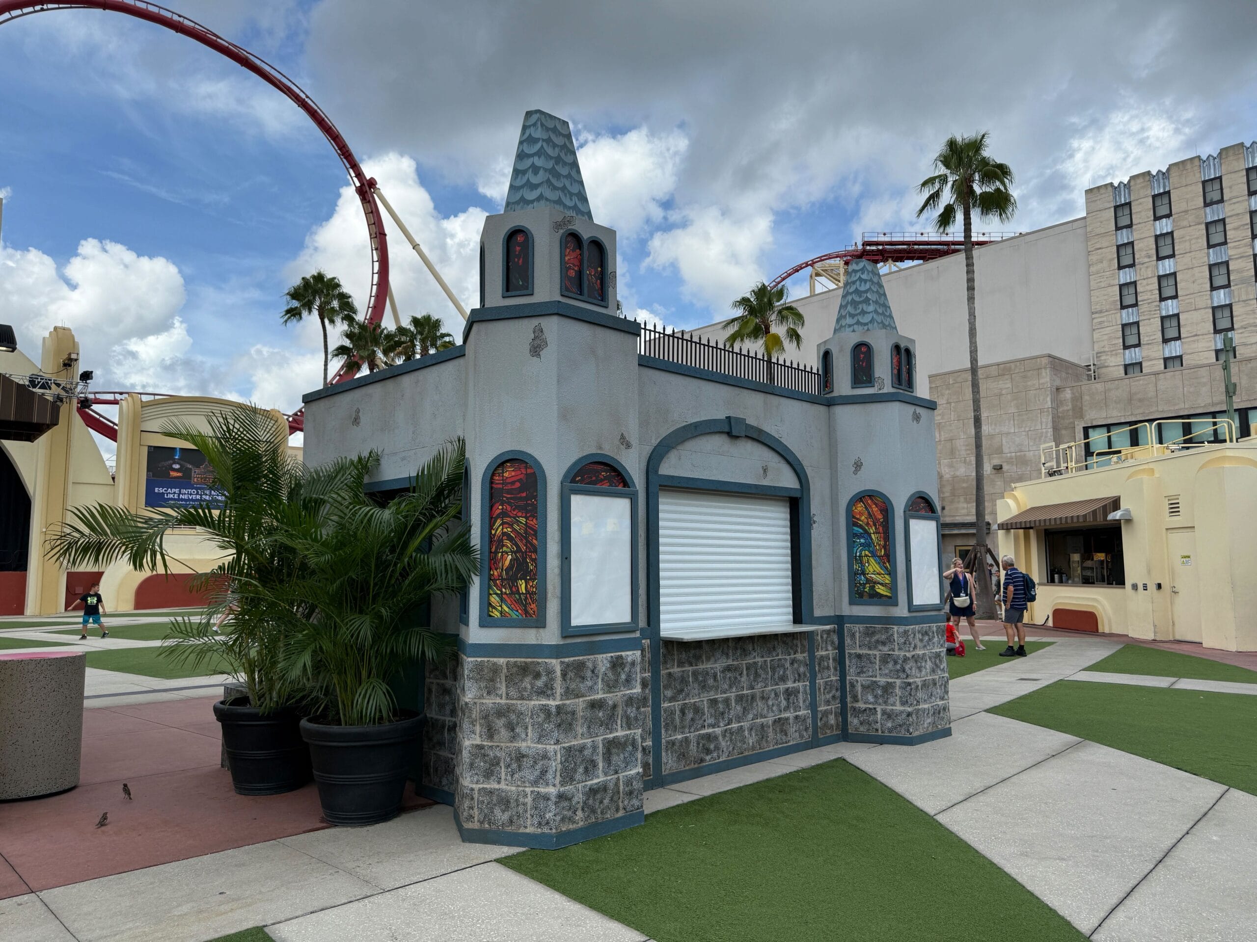 A small, castle-themed kiosk with closed shutters stands near a Firefly Fun House Sign by roller coaster tracks on a sunny day.