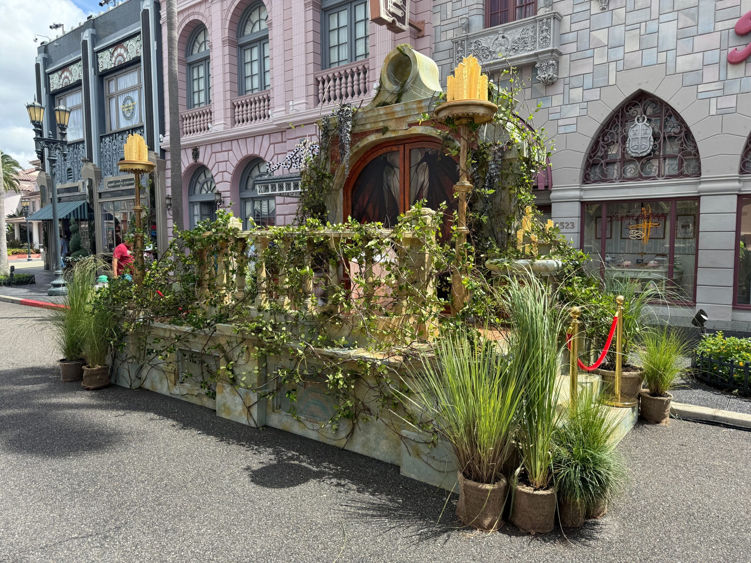 A parade float with greenery and vines sits on a city street, featuring a Firefly Fun House Sign in front of ornate buildings.