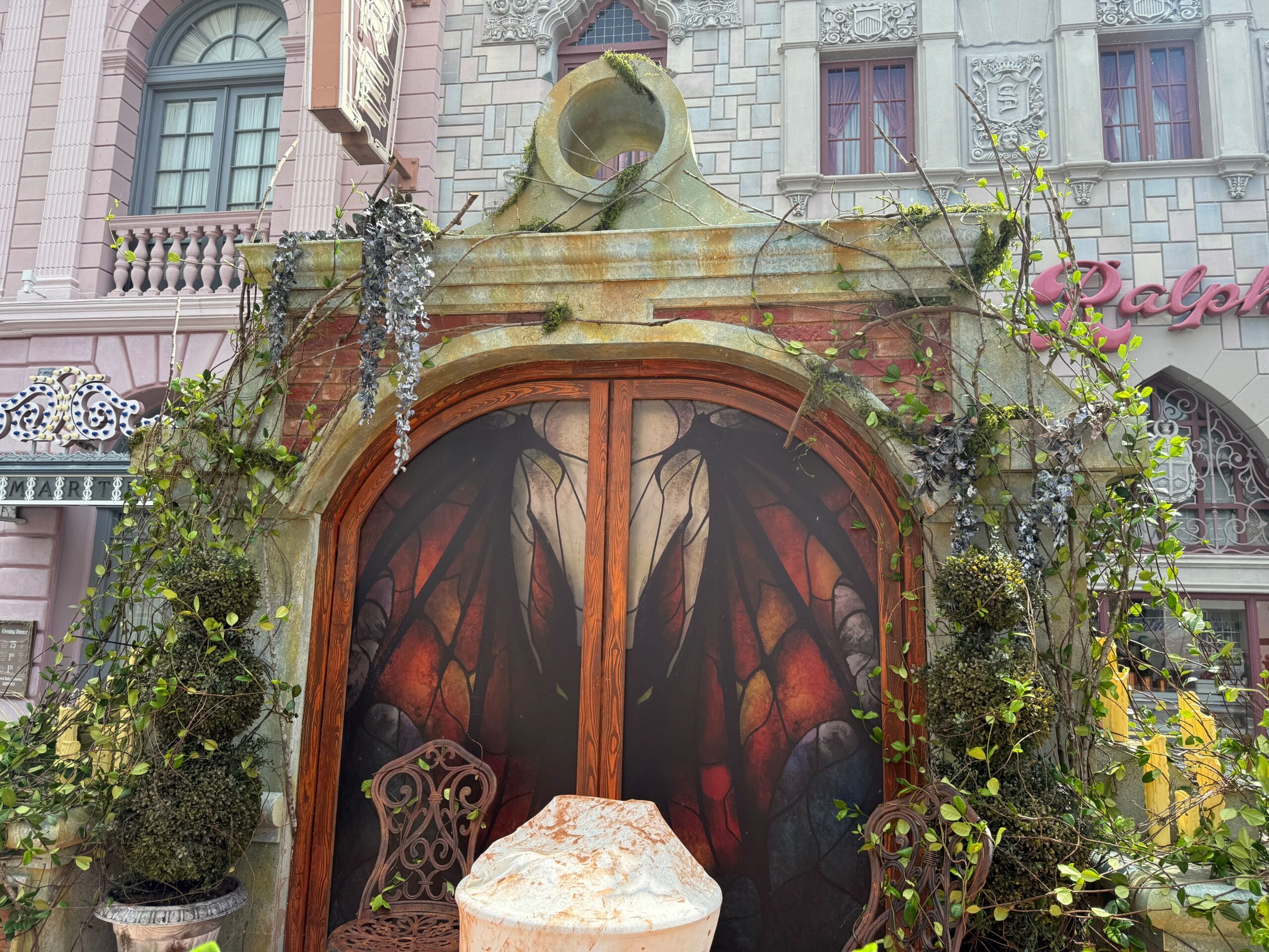 Decorative arched doorway with vine-covered stone, a stained-glass bat wing, and Firefly Fun House Sign on a weathered table.