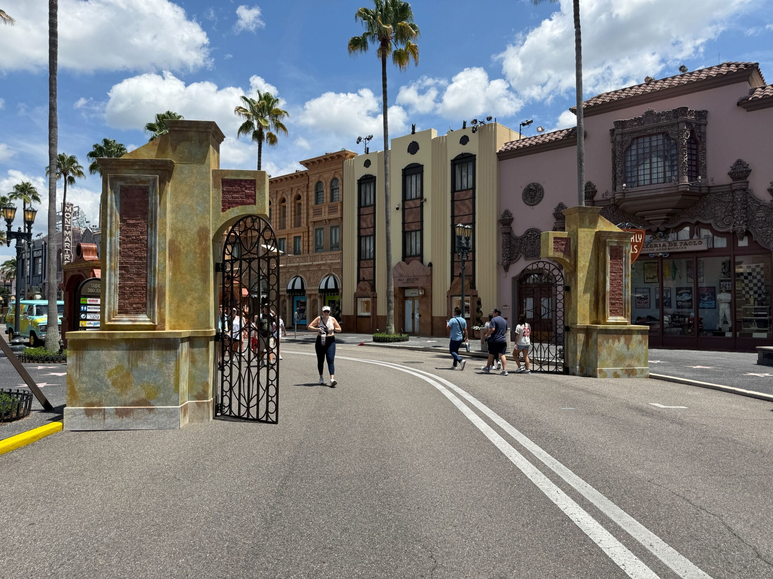 Arched gates open to a street lined with people and buildings under a sunny, partly cloudy sky.