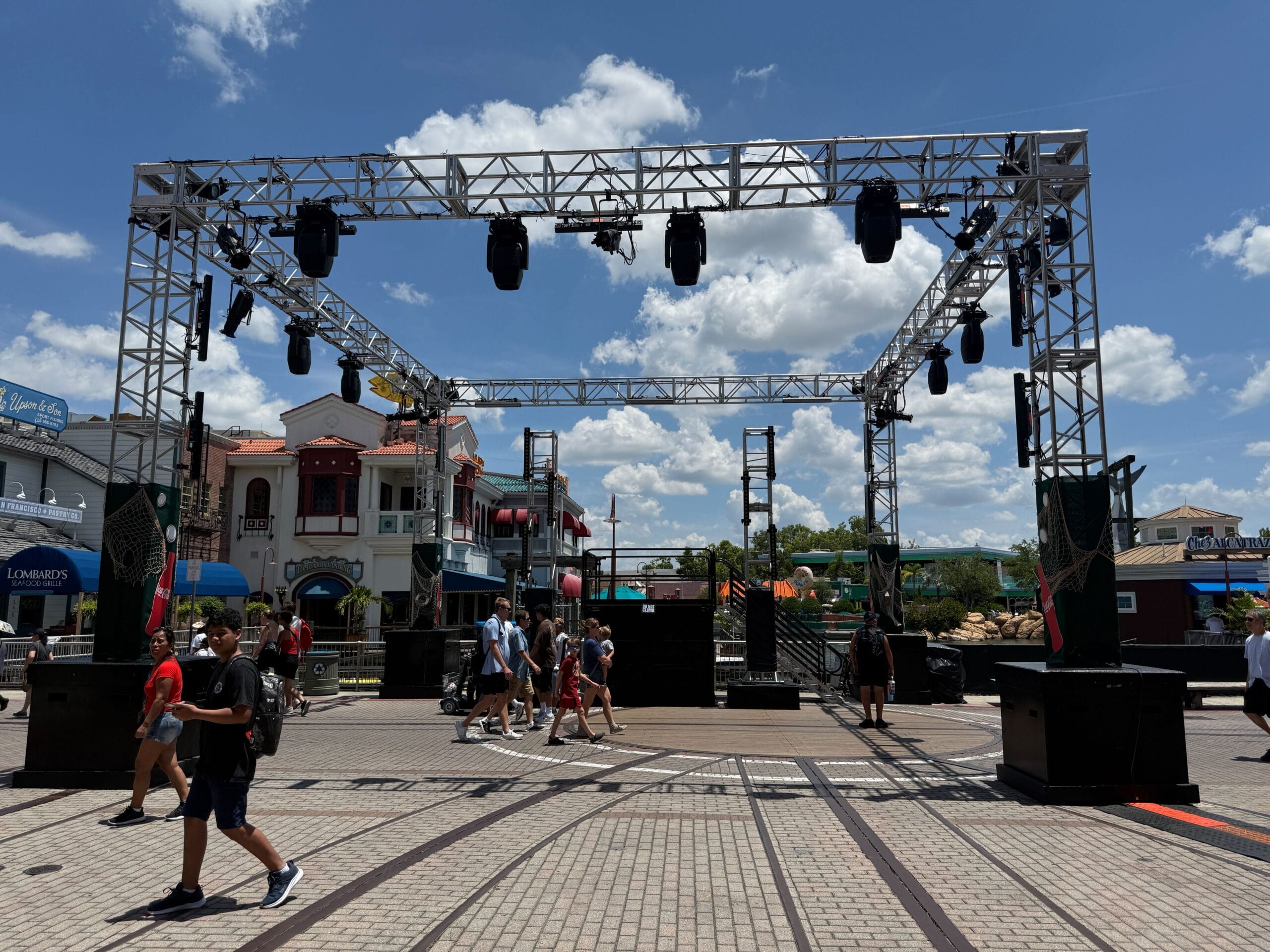 People walk under a large outdoor stage setup with lights in a lively plaza on a sunny day.
