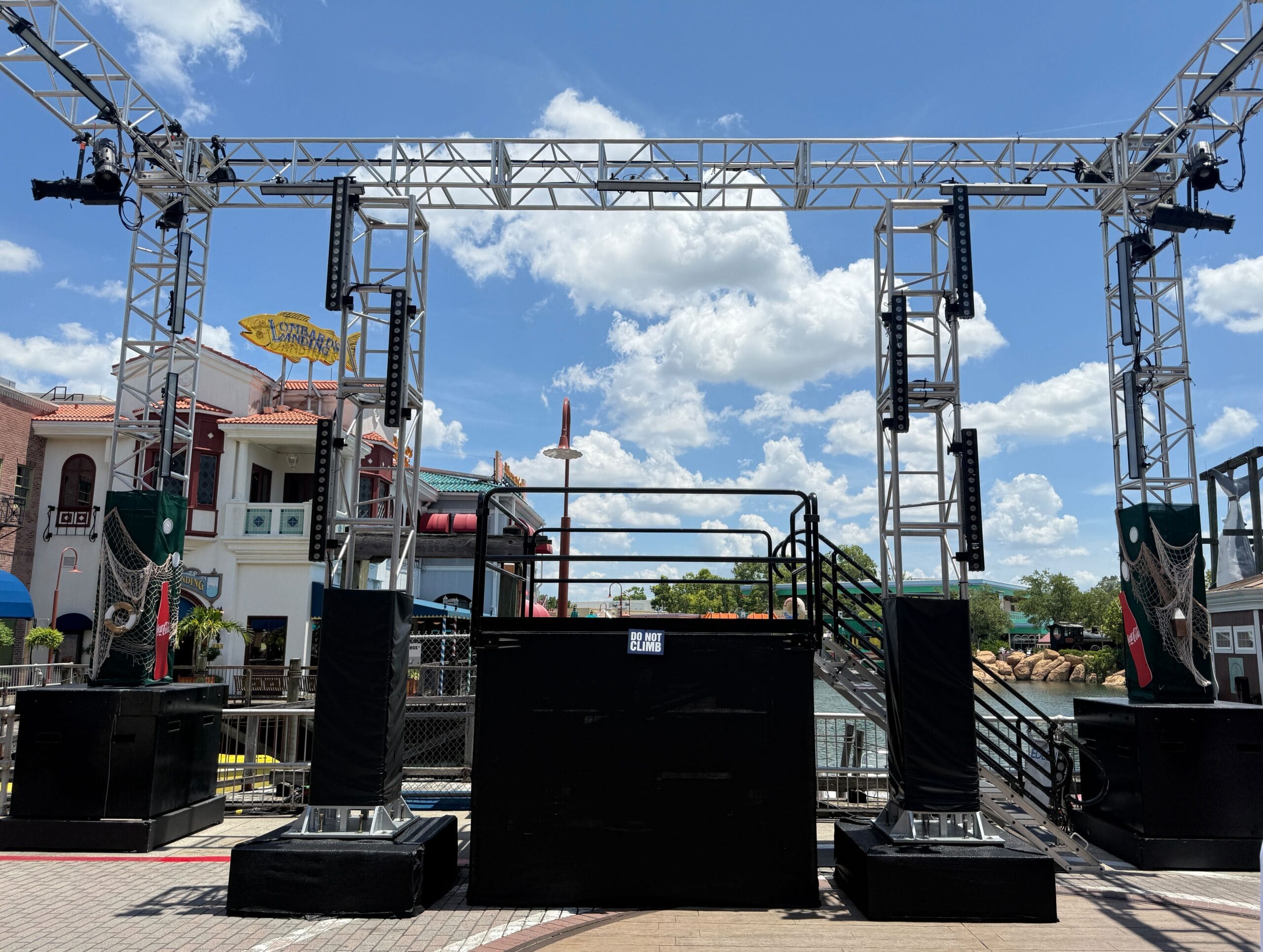 A metal stage structure with speakers stands outdoors near water, with buildings and a blue sky with clouds in the background.