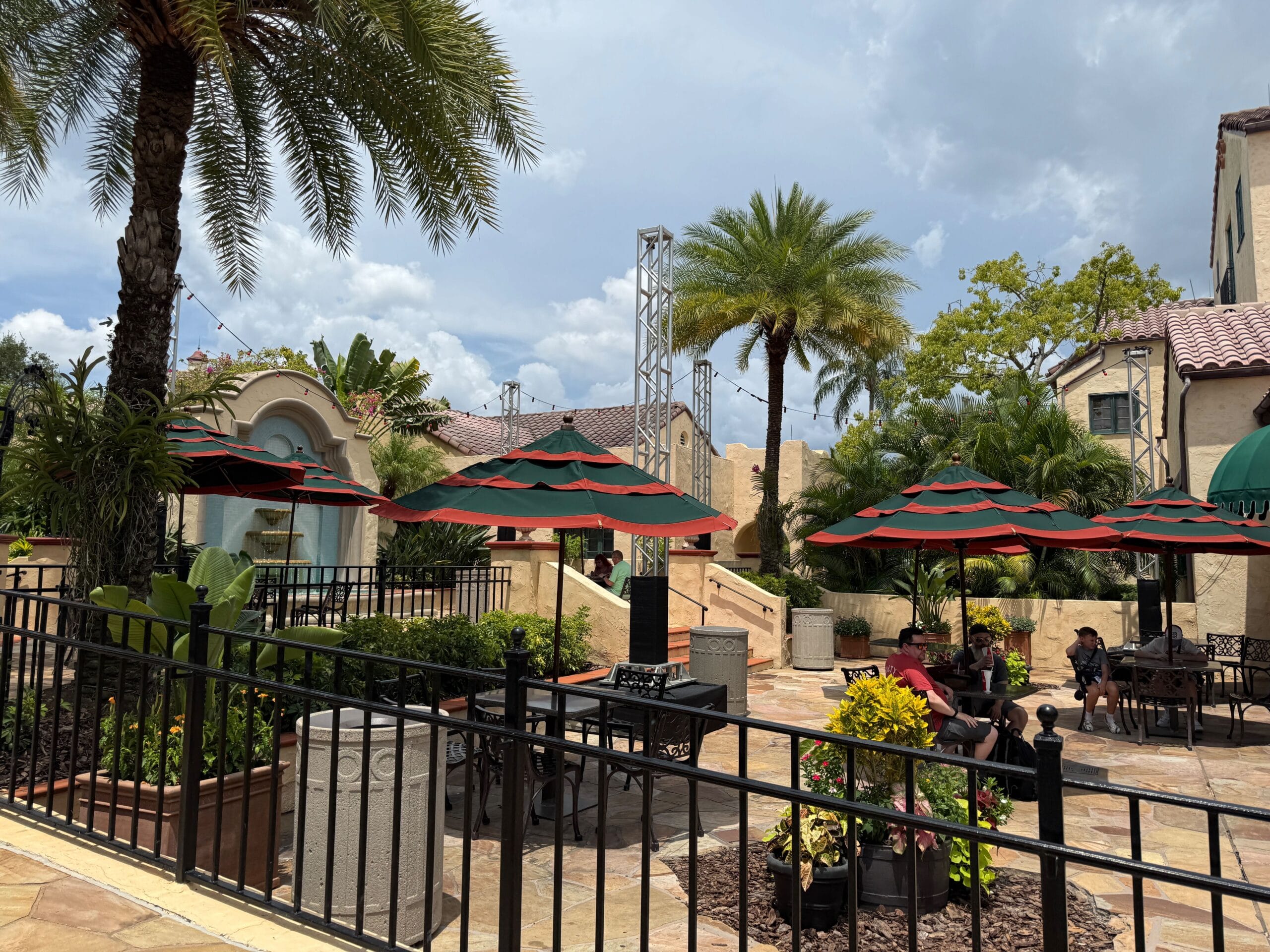 Outdoor patio with red and green umbrellas, palm trees, and people sitting at tables on a cloudy day.