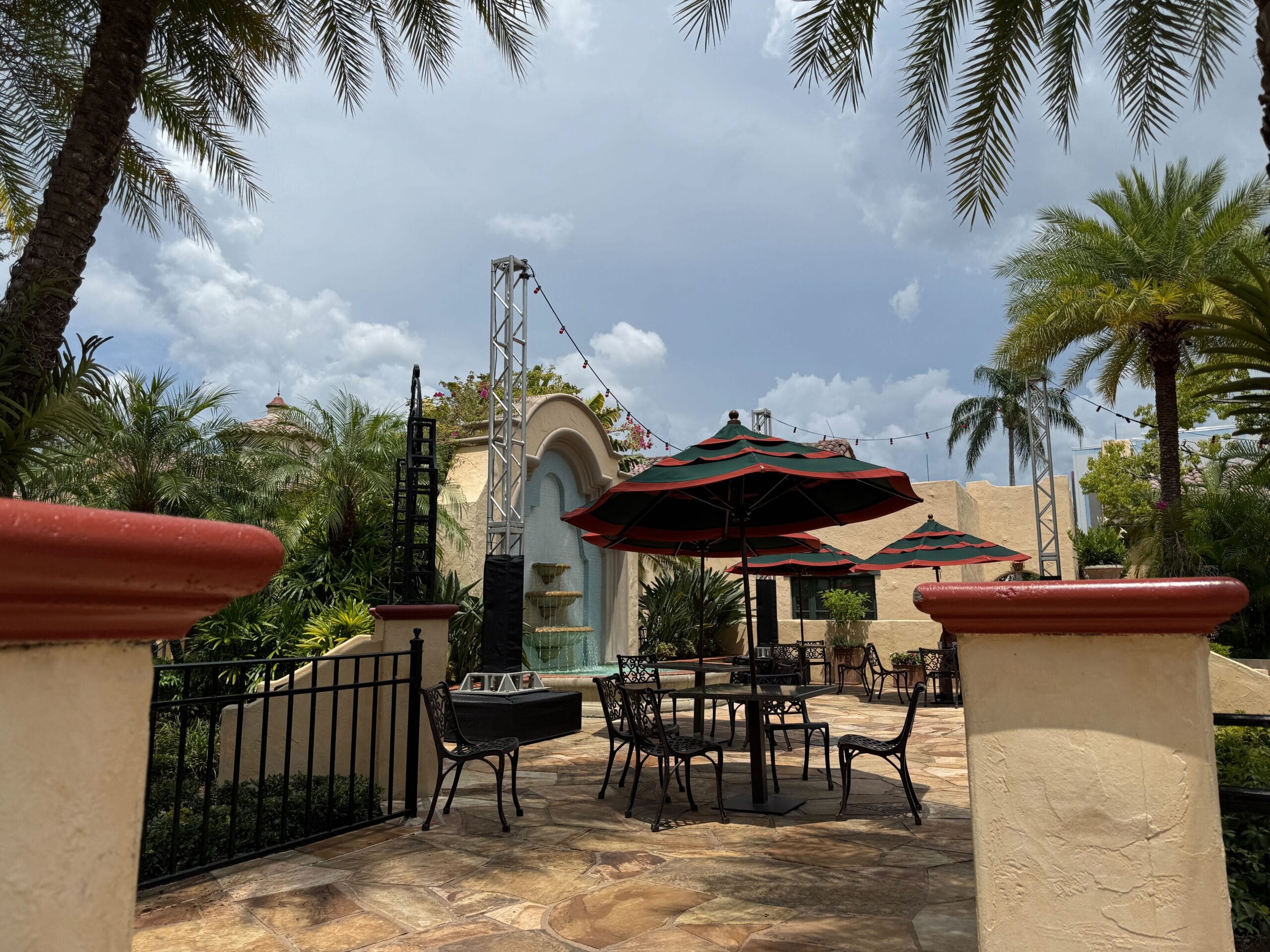 Outdoor patio with black tables, chairs, and umbrellas surrounded by palm trees and a decorative fountain under a partly cloudy sky.