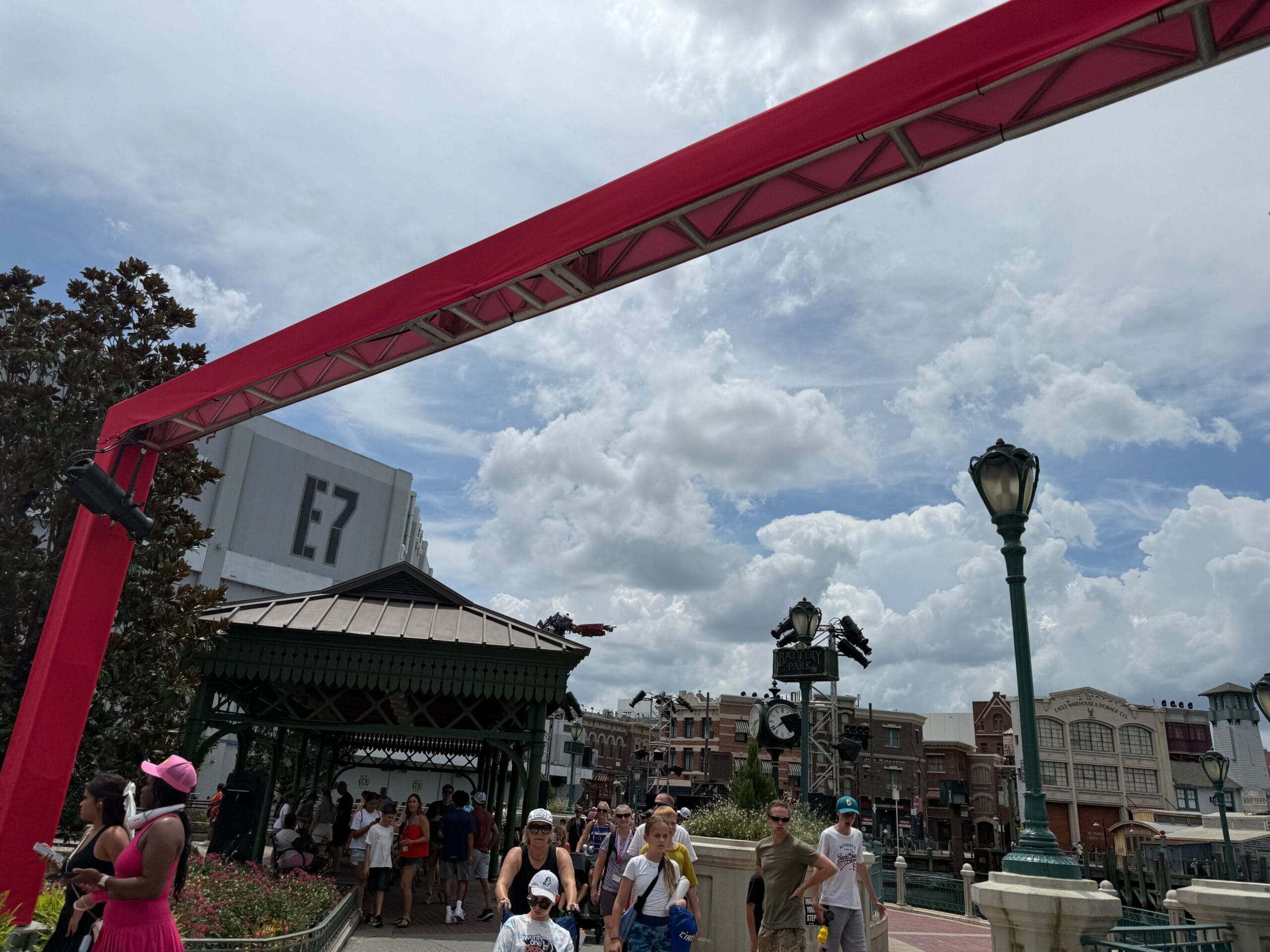 People walking outdoors under a red truss with buildings, lamppost, and a cloudy sky in the background.
