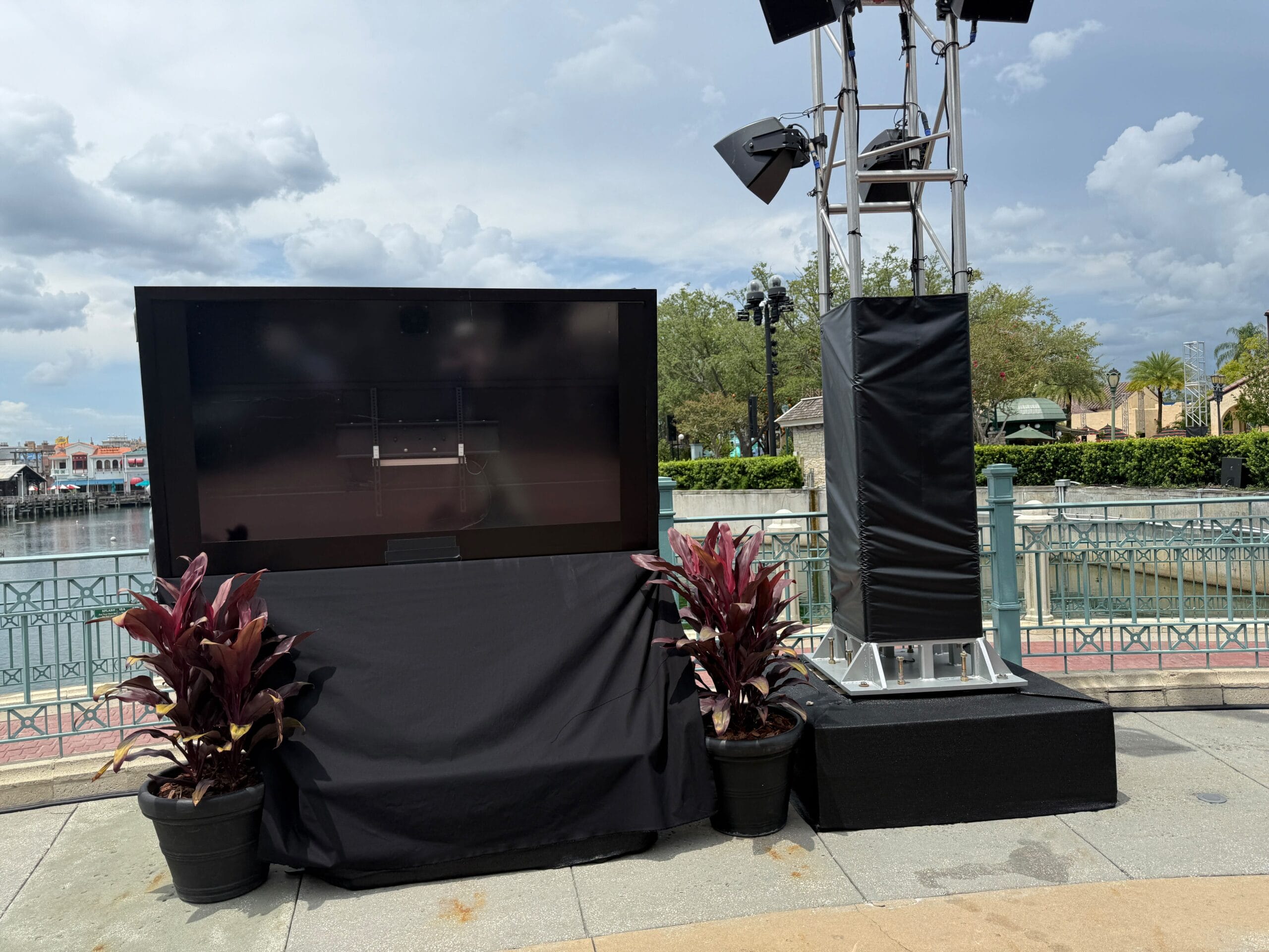 A large outdoor TV screen and lighting rig, surrounded by potted plants, set up by a waterfront on a cloudy day.