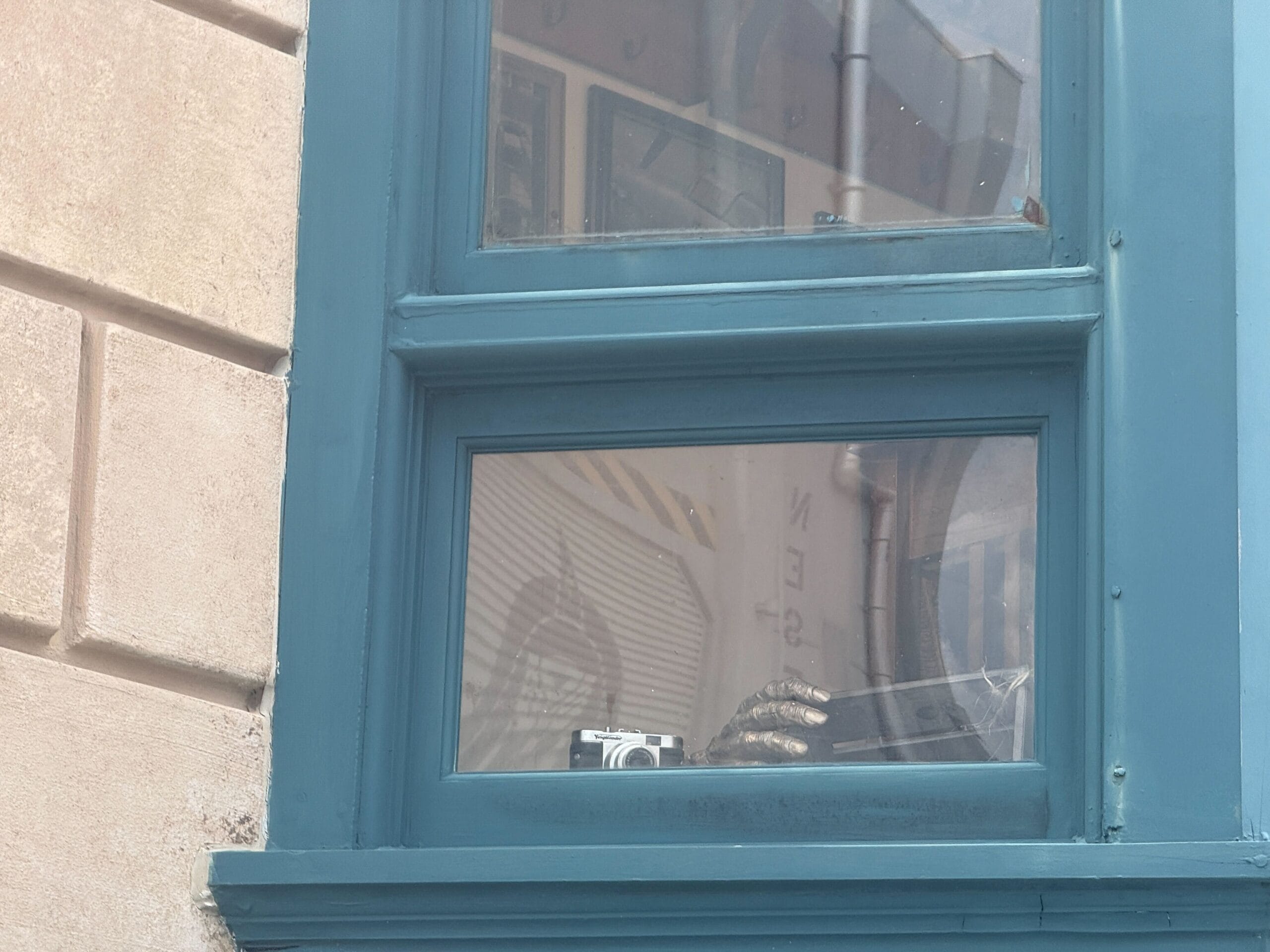 A silver metallic hand and a camera are visible through a blue-framed window from the outside.