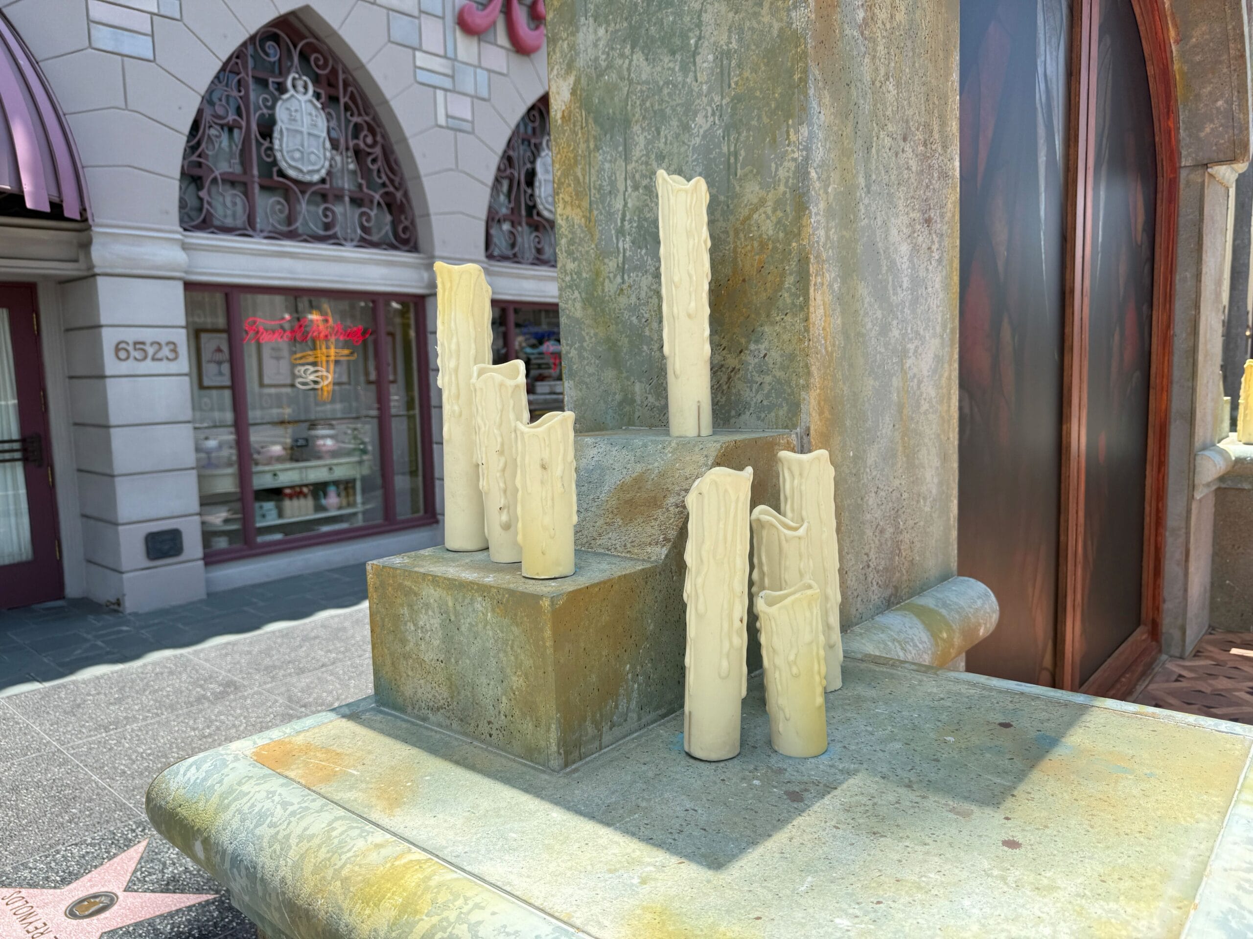 Seven decorative, faux wax candles displayed on a stone pedestal outside a building on a sunny day.
