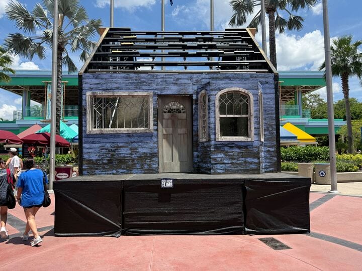 A blue, weathered house facade is displayed on a platform outdoors under a sunny sky with palm trees nearby.