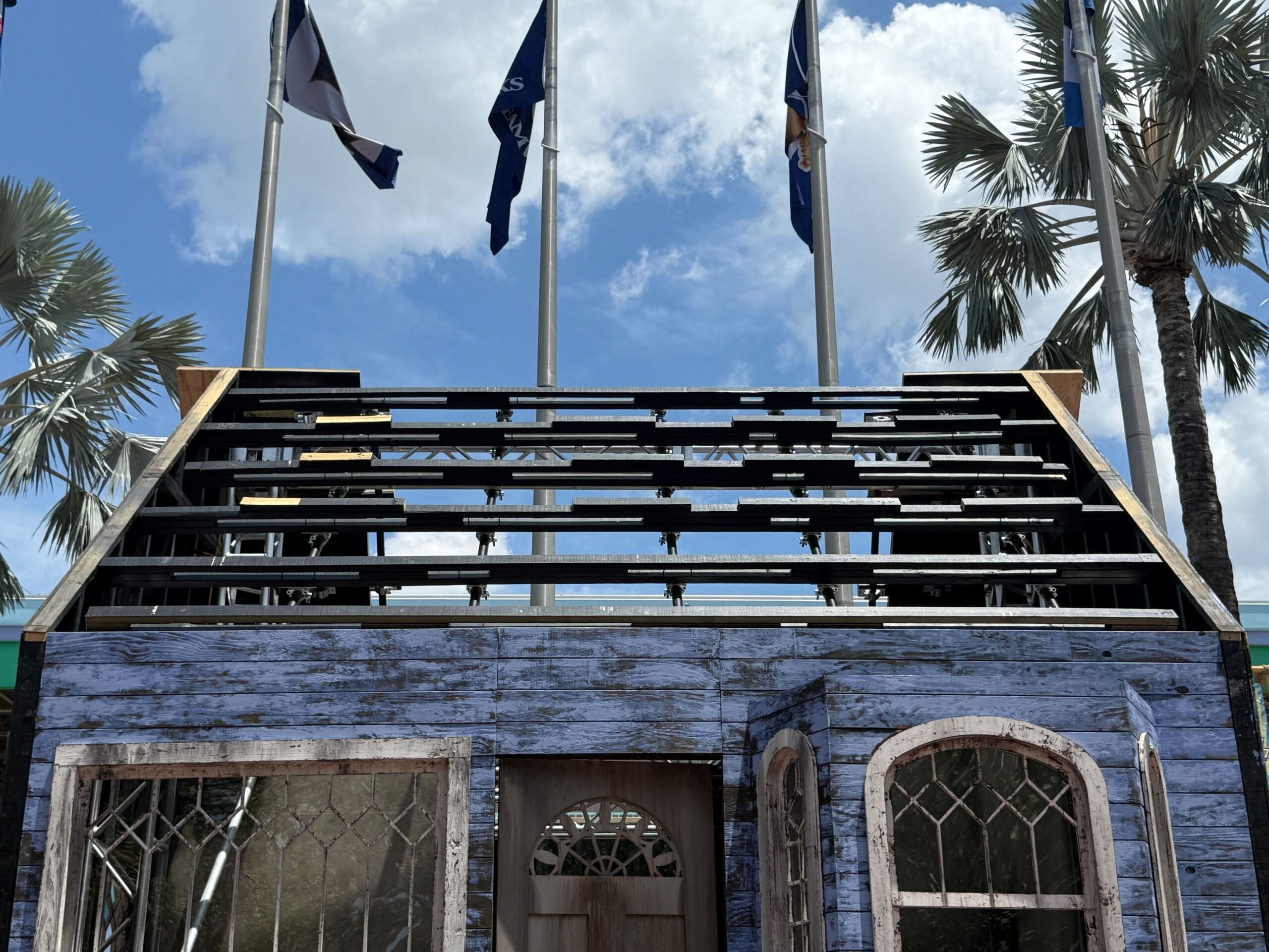 A weathered blue wooden house facade with missing roof, flags, and palm trees in the background under a partly cloudy sky.