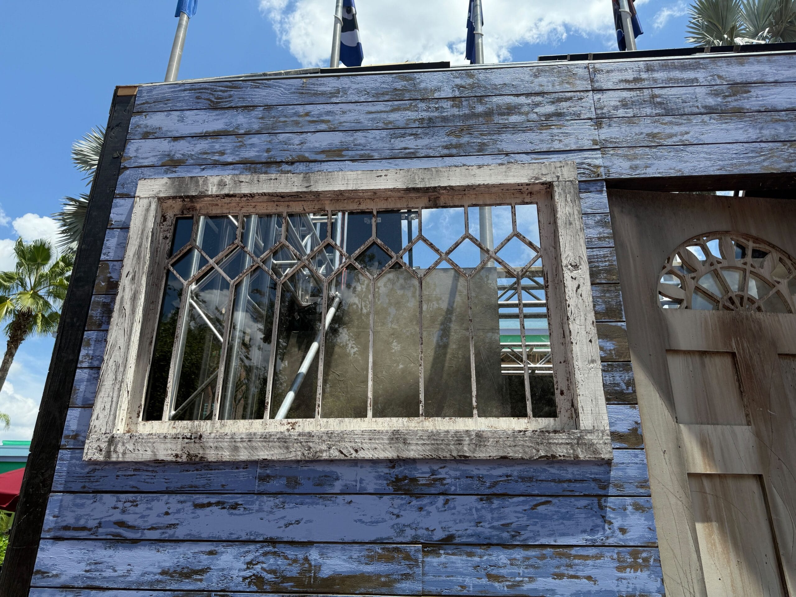 Weathered blue wooden wall with a rectangular window featuring diamond-patterned panes under a sunny sky.