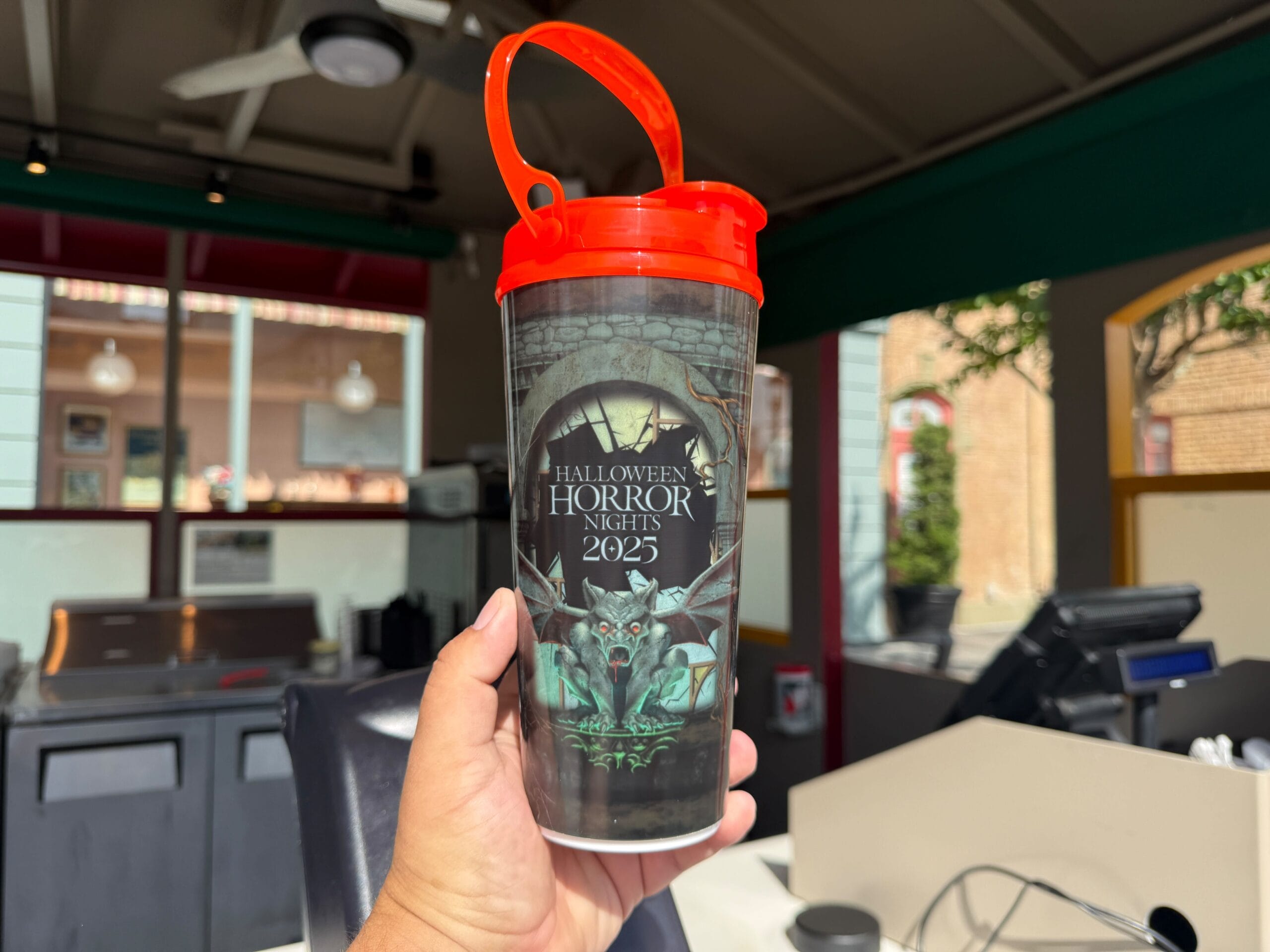 A hand holds a 2025 Halloween Horror Nights cup with a red lid and spooky design inside a concession stand.