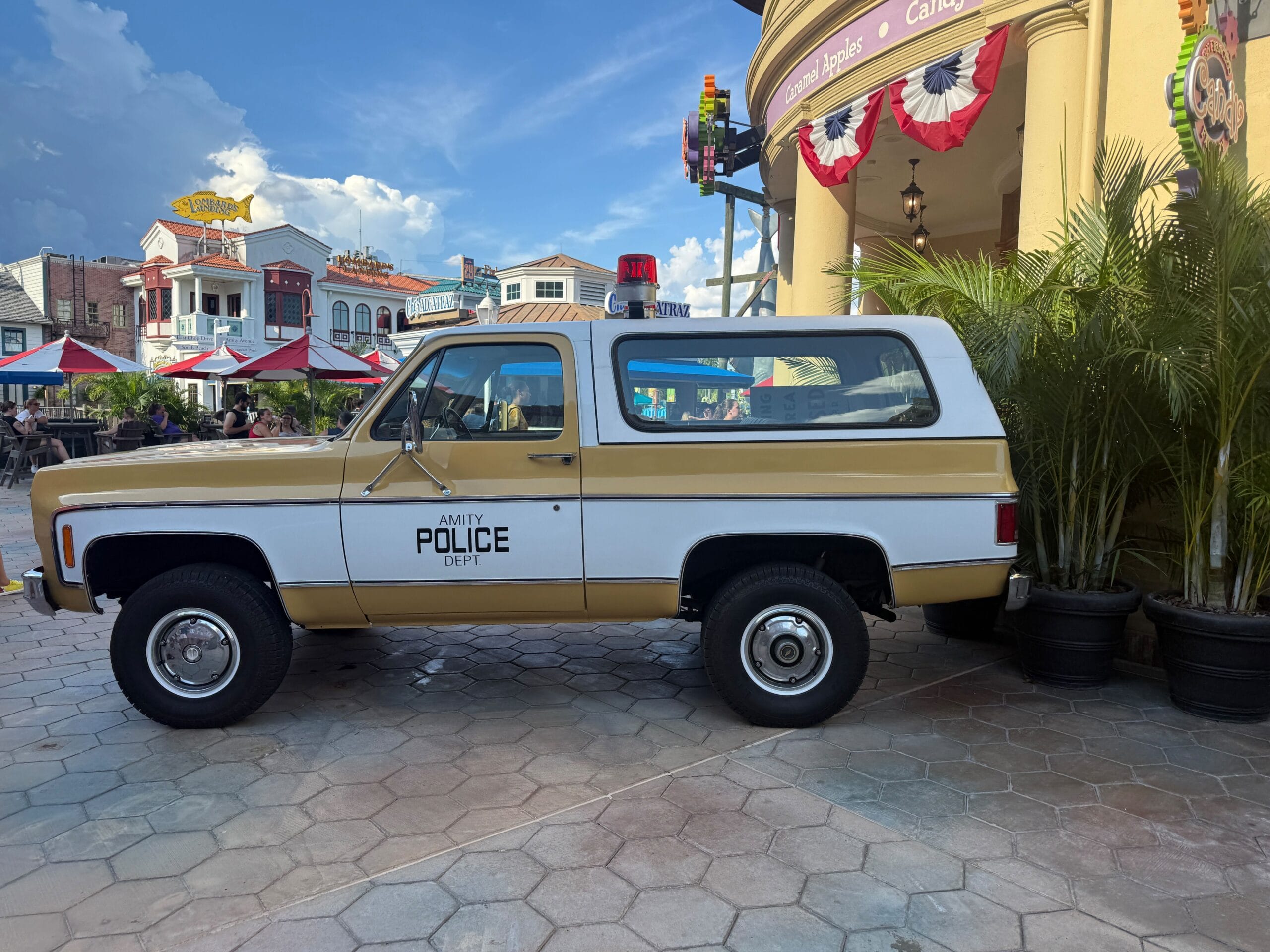 A vintage Amity Police Dept. SUV parked by a building with palm plants and festive decorations.