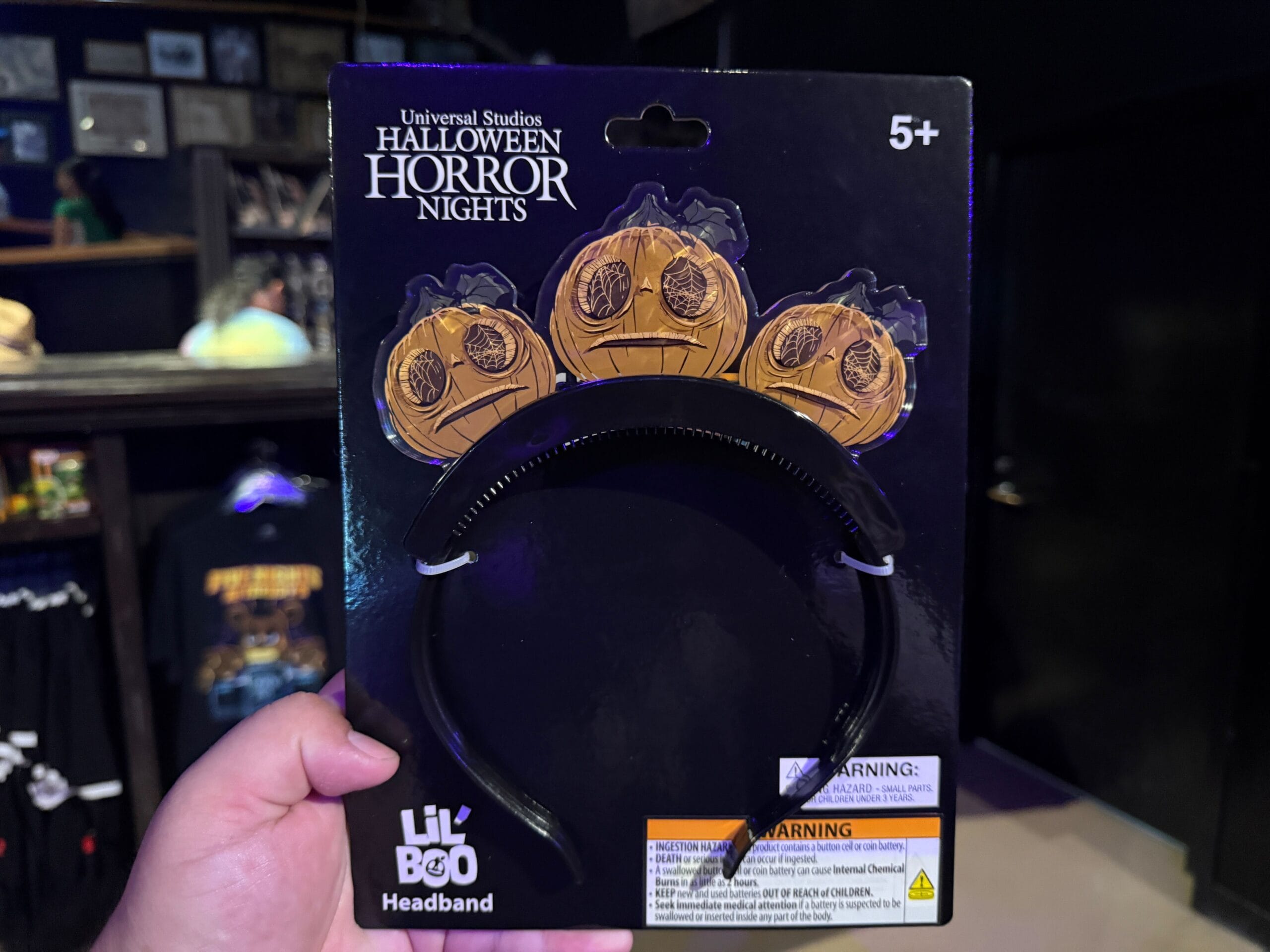 A hand holds a Halloween Horror Nights headband with three pumpkin decorations labeled "Lil Boo Headband.